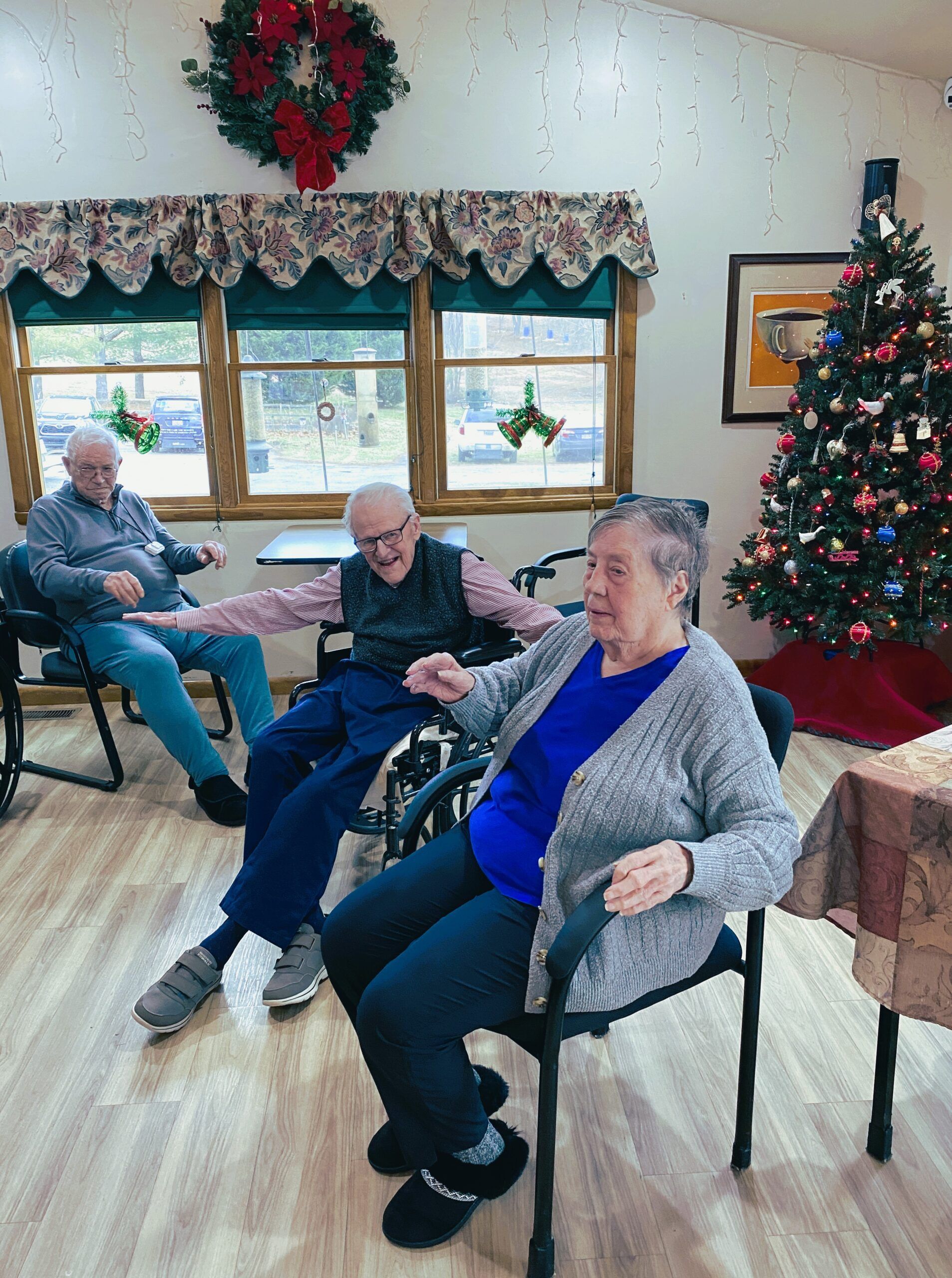 Three people seated in a room decorated for Christmas with a large tree, wreaths, and festive decorations.