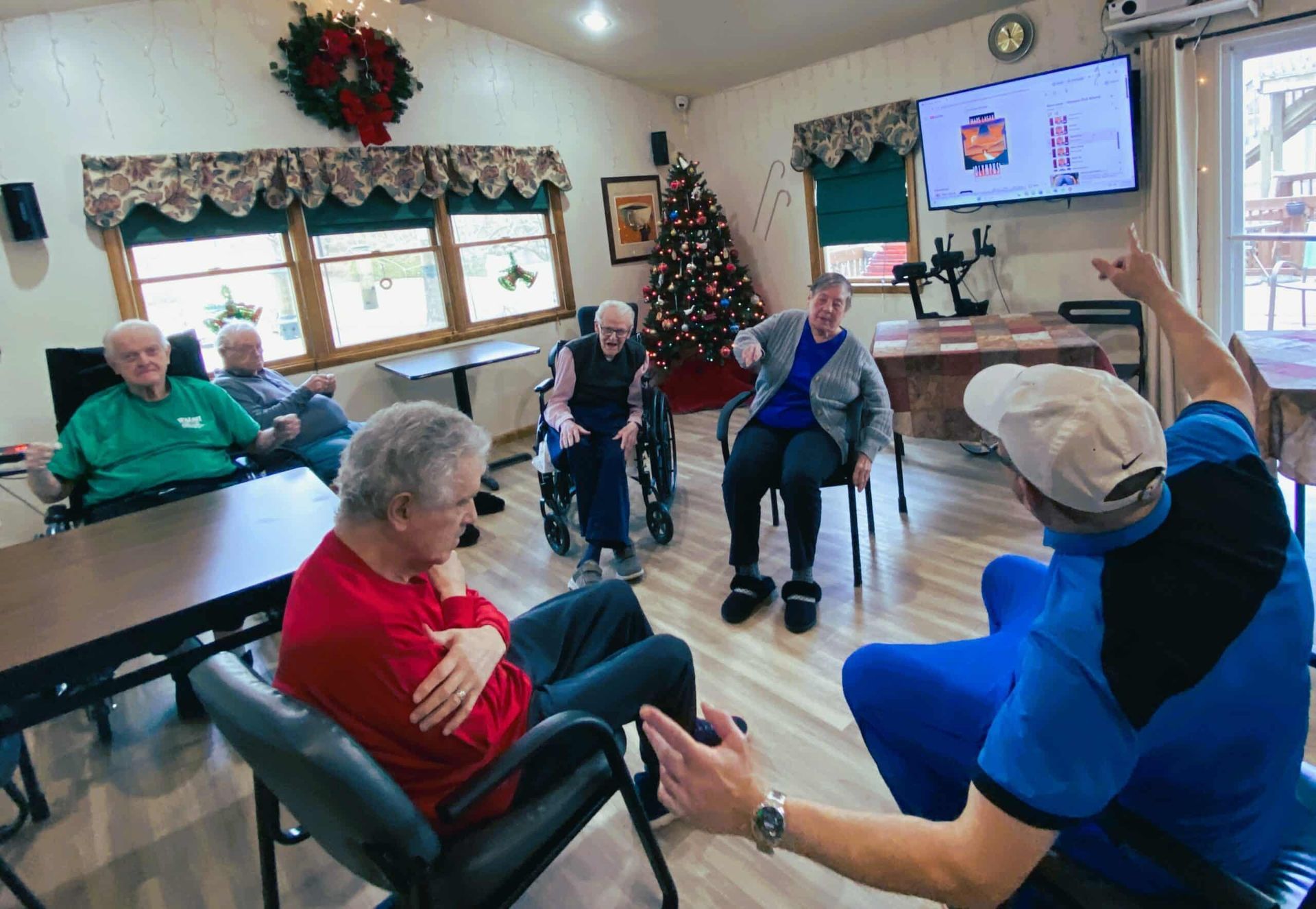 A group gathers in a room with a Christmas tree; a person points at a screen while others sit and watch.