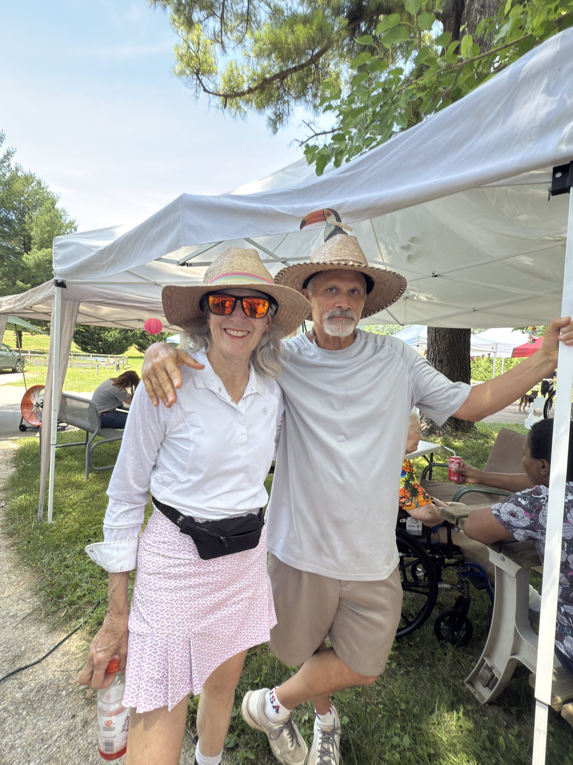 Two people wearing straw hats and sunglasses stand smiling under a white canopy at an outdoor gathering.