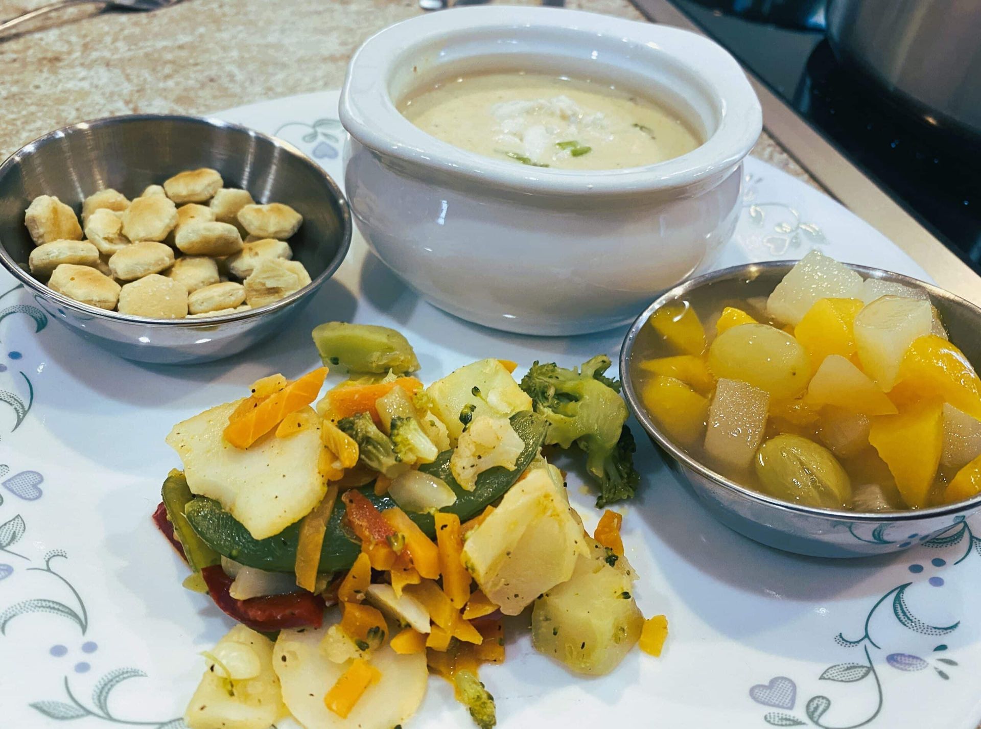 A meal featuring a bowl of creamy soup, a small bowl of crackers, a cup of fruit, and a vegetable stir-fry on a plate.