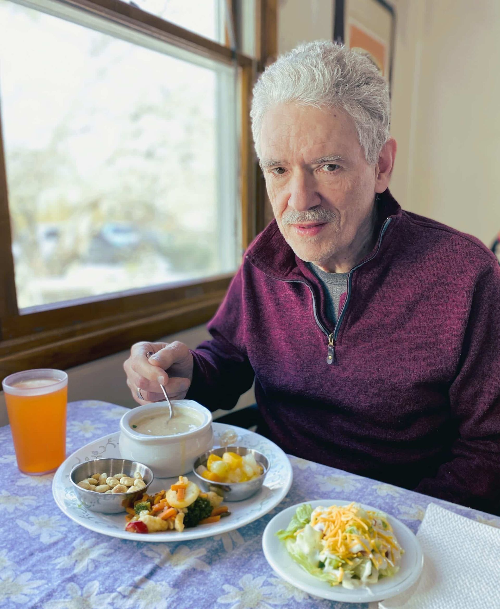 An individual sits at a table near a window, eating a meal of soup, salad, and sides from a plate and a separate bowl.