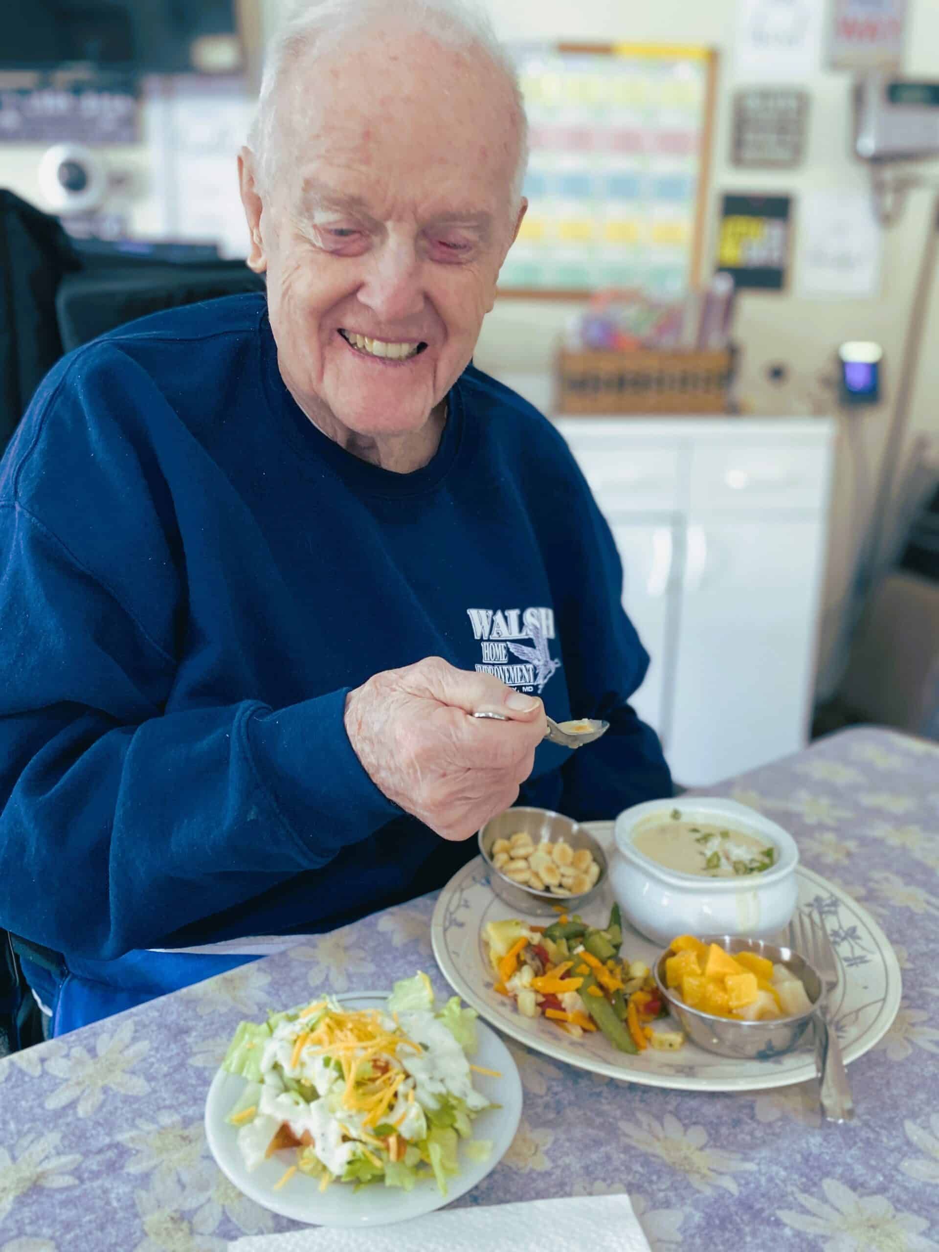 A smiling person in a blue sweatshirt sits at a table and eats a meal with a spoon, accompanied by soup, salad, and fruit.