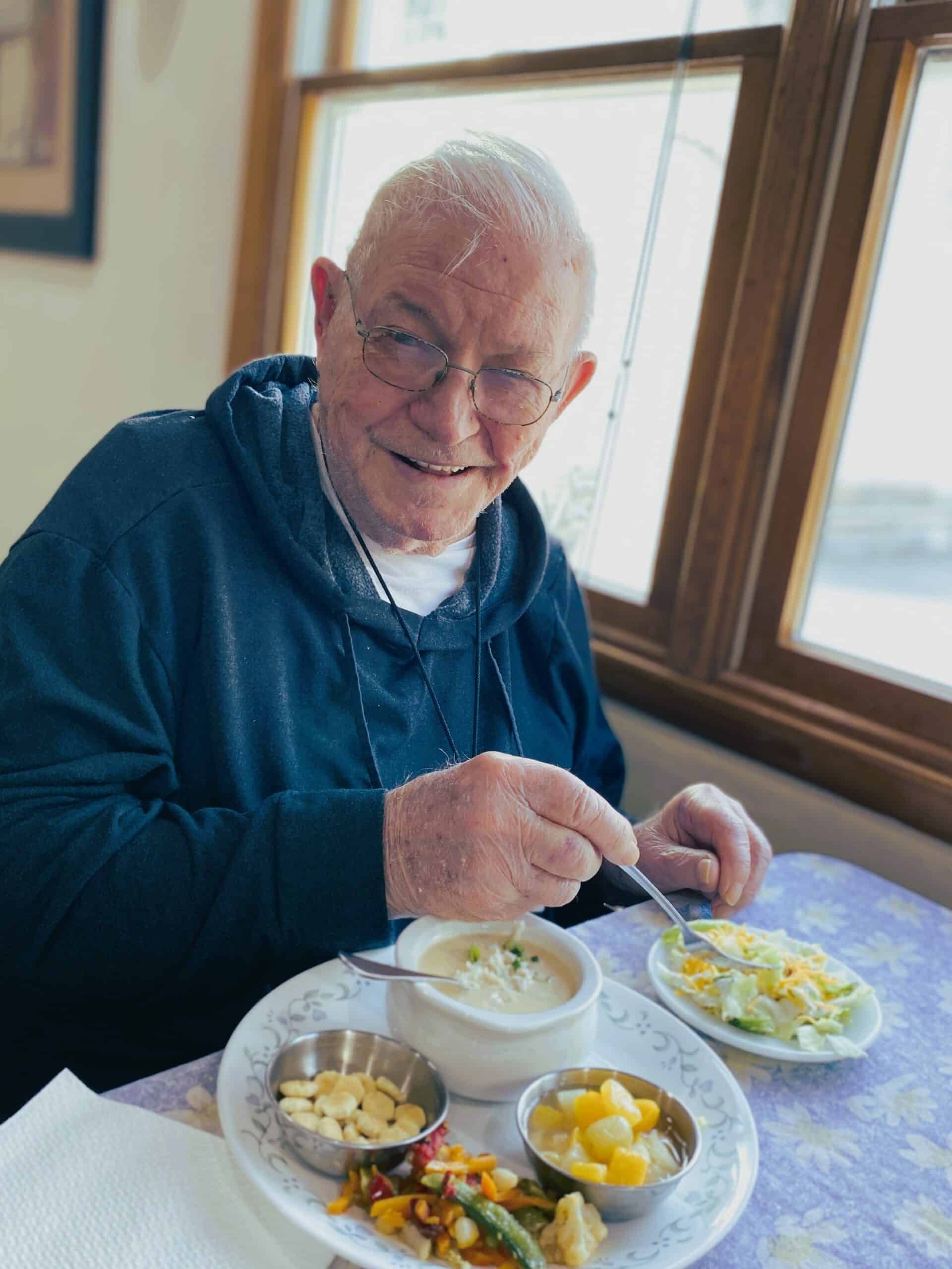 A smiling person in a blue hoodie eating a meal of soup, salad, crackers, and fruit at a table by a window.