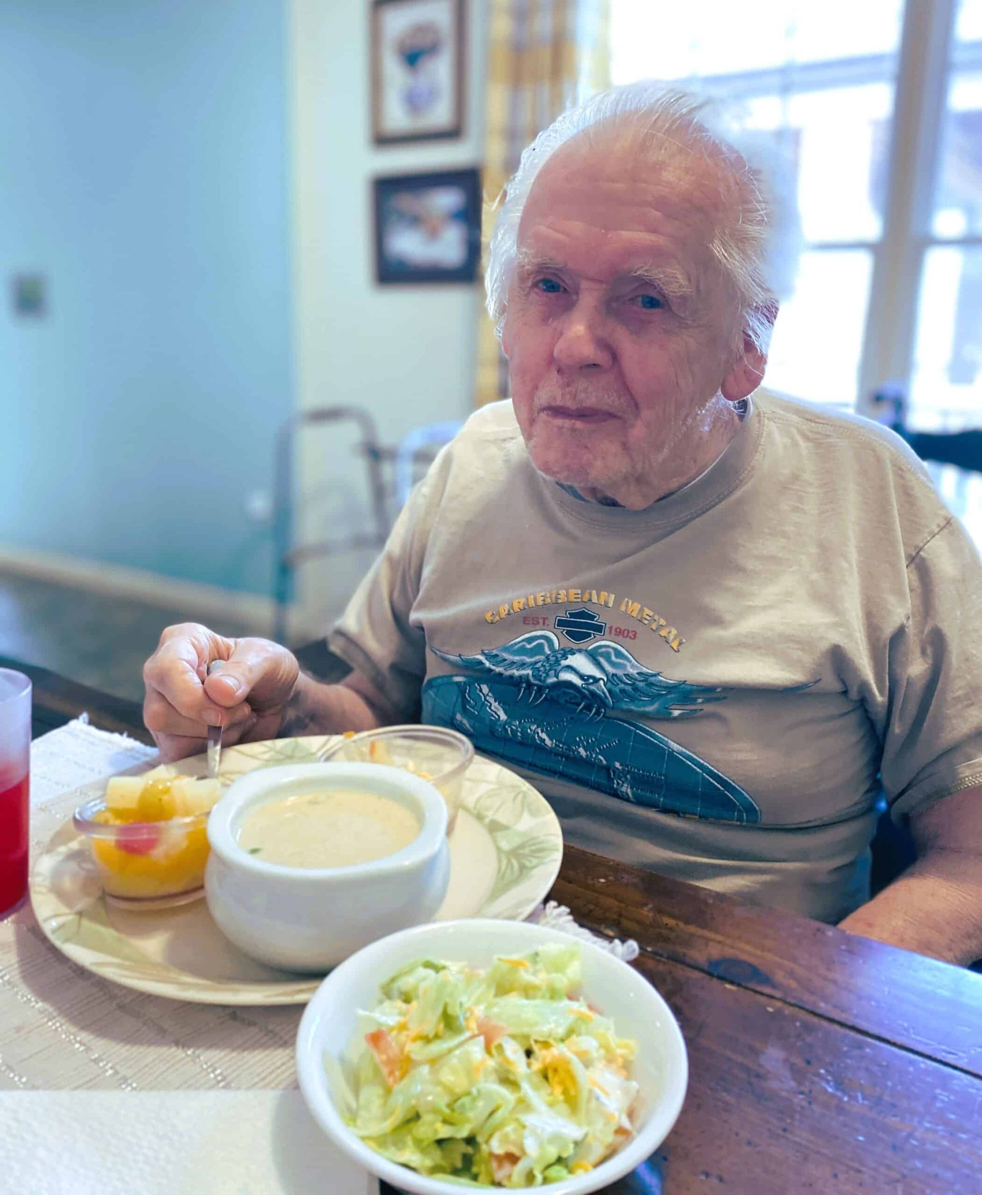 An individual sitting at a table with a meal of soup, fruit, and coleslaw, looking toward the camera.