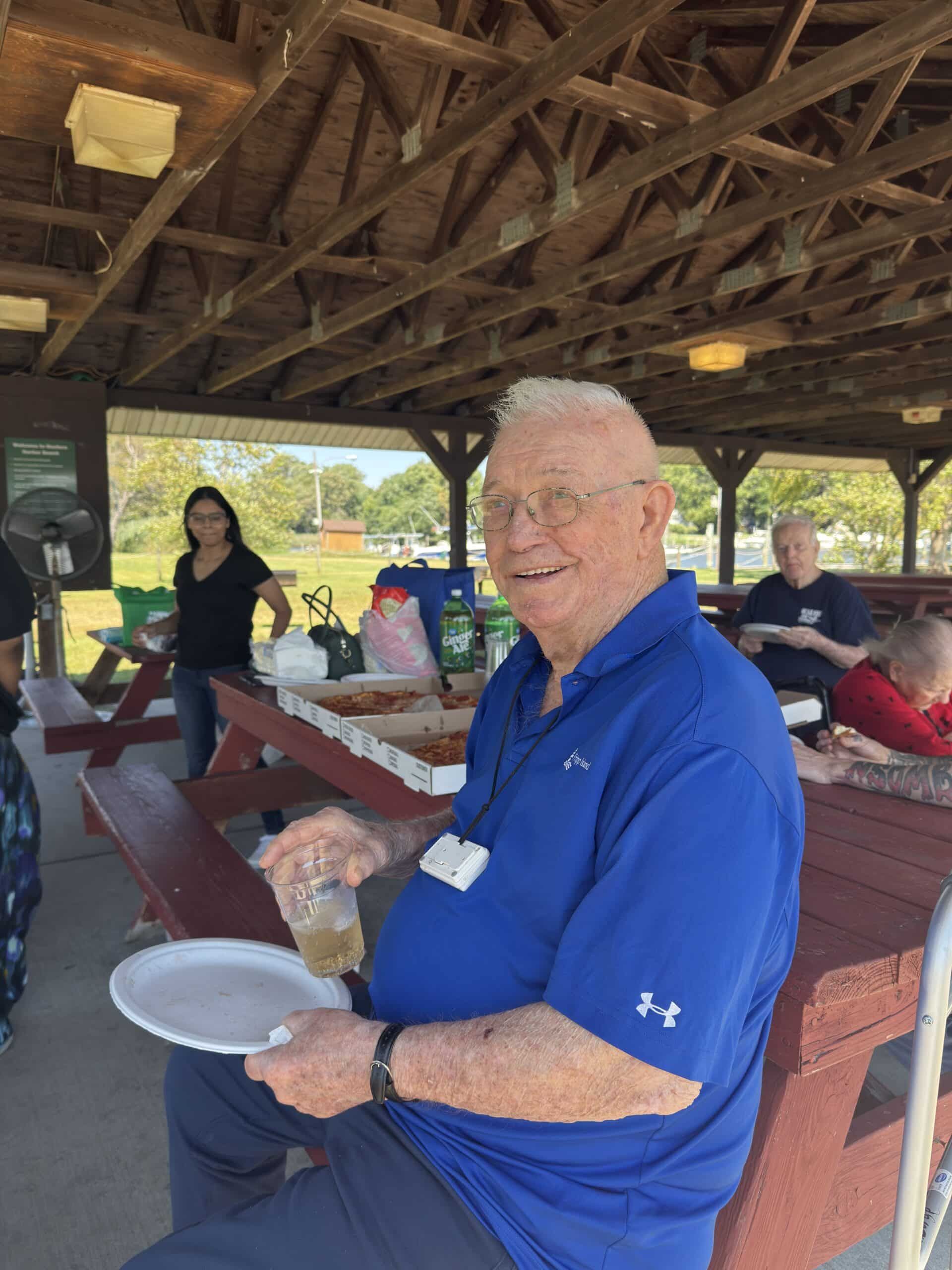 A smiling person in a blue polo shirt holds a plate and drink under a wooden pavilion at an outdoor gathering.