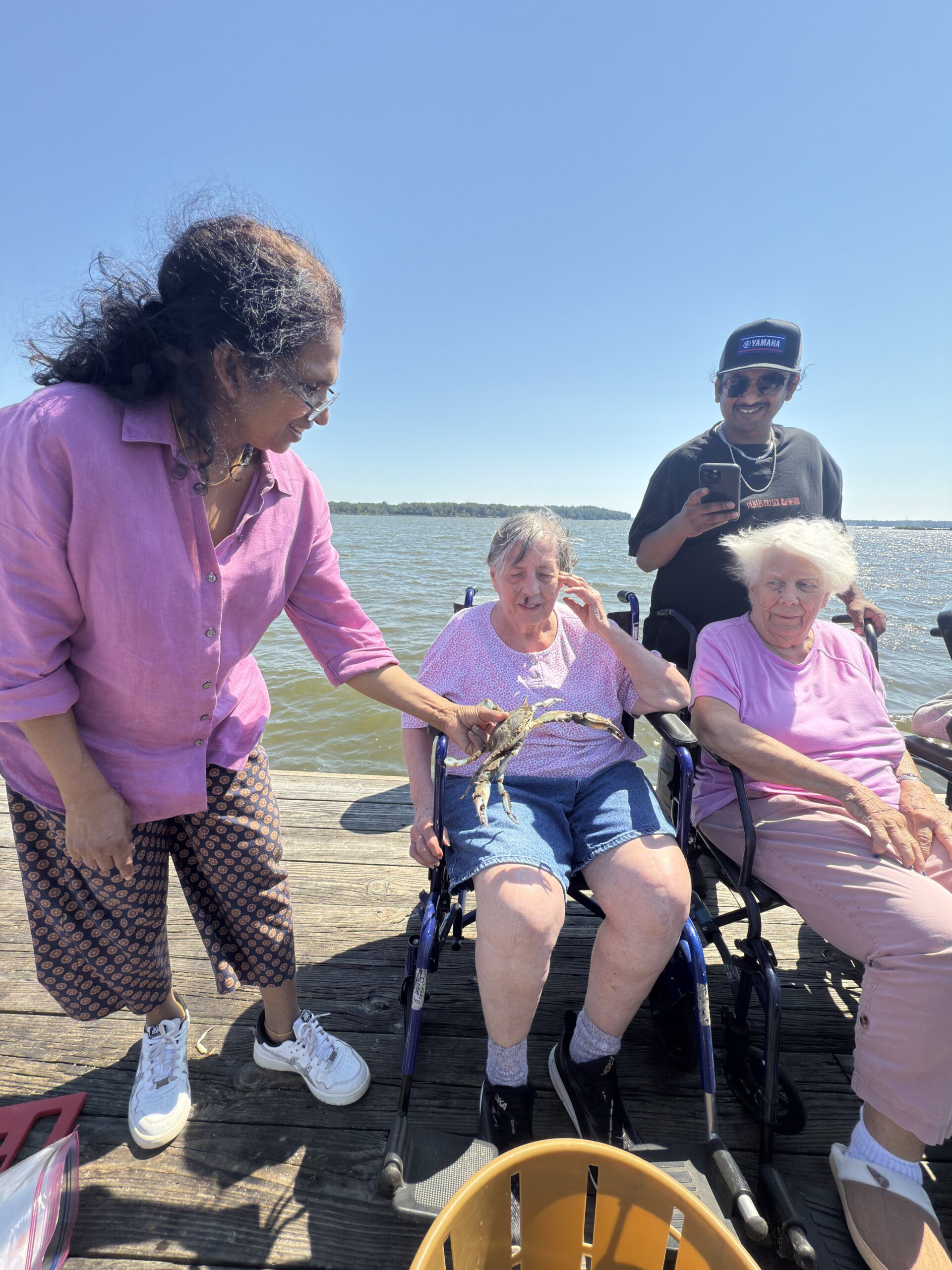 Four people gather on a wooden pier by the water; two individuals in wheelchairs are assisted by two others standing nearby.