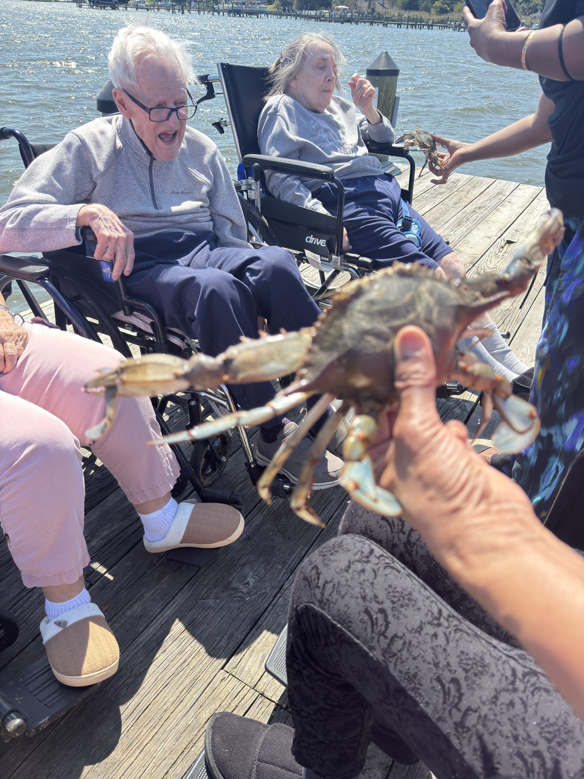 A person holds a live blue crab on a wooden dock in front of two individuals seated in wheelchairs.