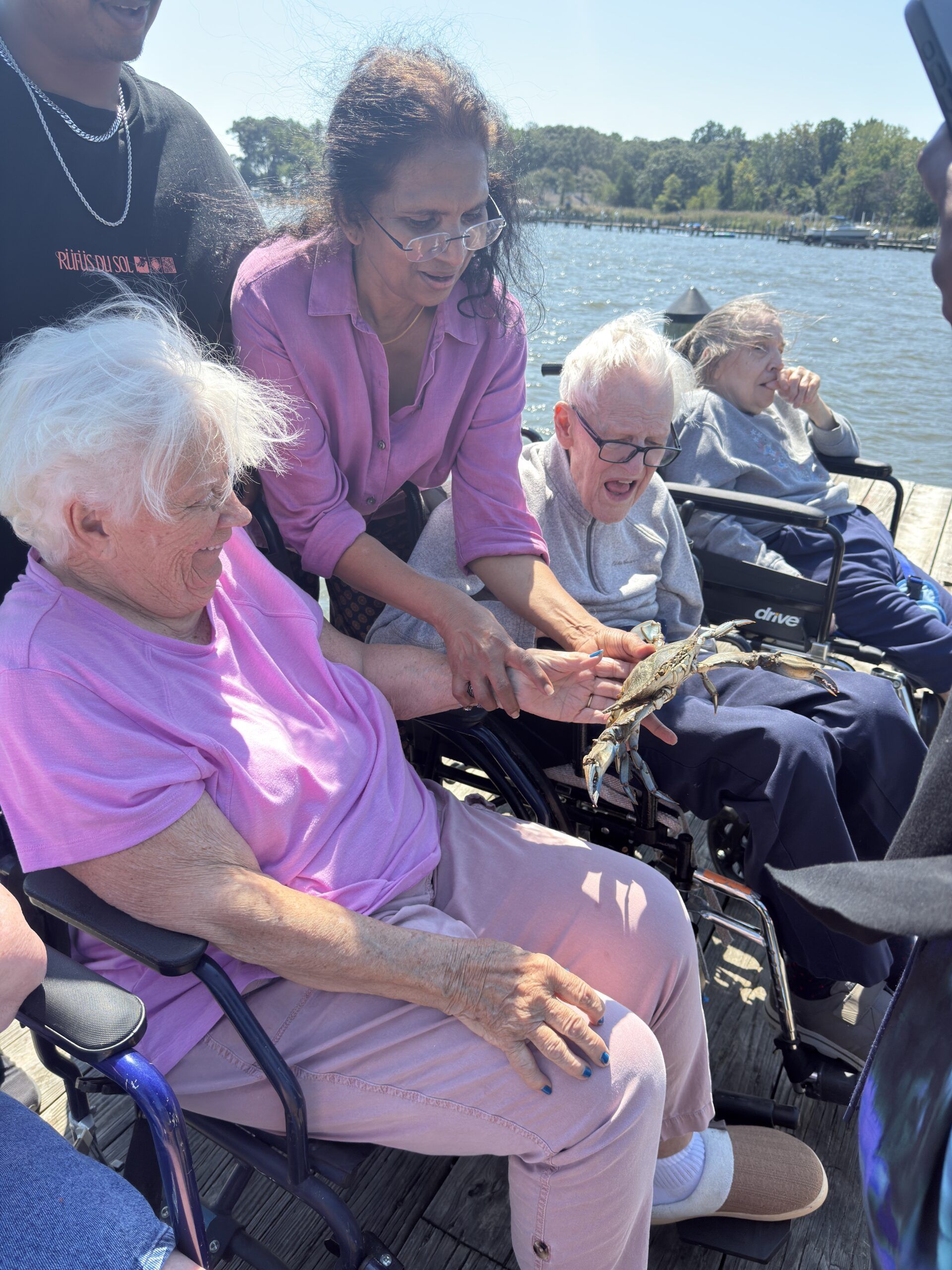 A woman stands behind two seated people by the water, showing them a crab in her hands on a sunny day.