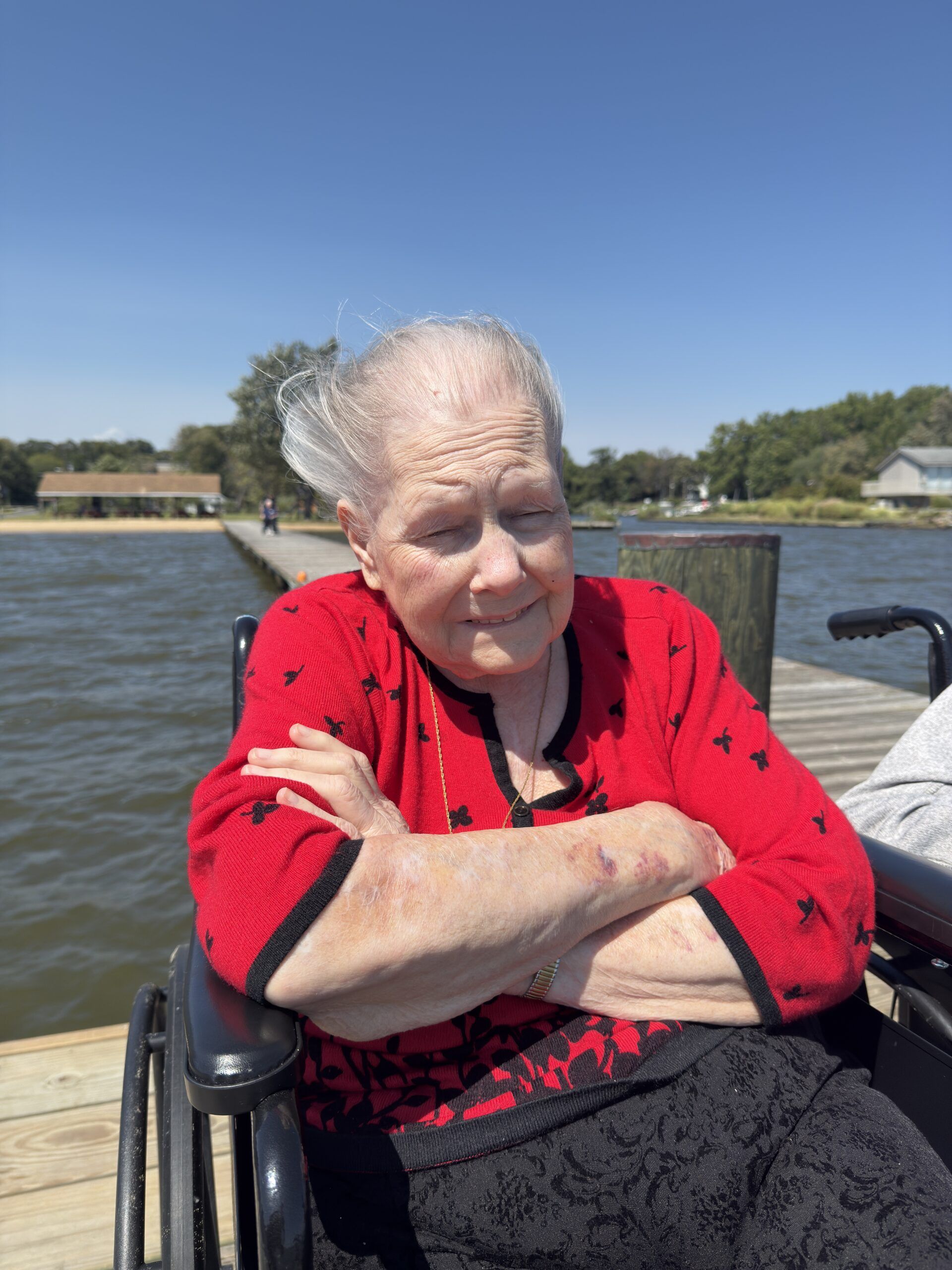 A person sitting in a wheelchair on a wooden pier by a body of water, wearing a red patterned top with arms crossed.