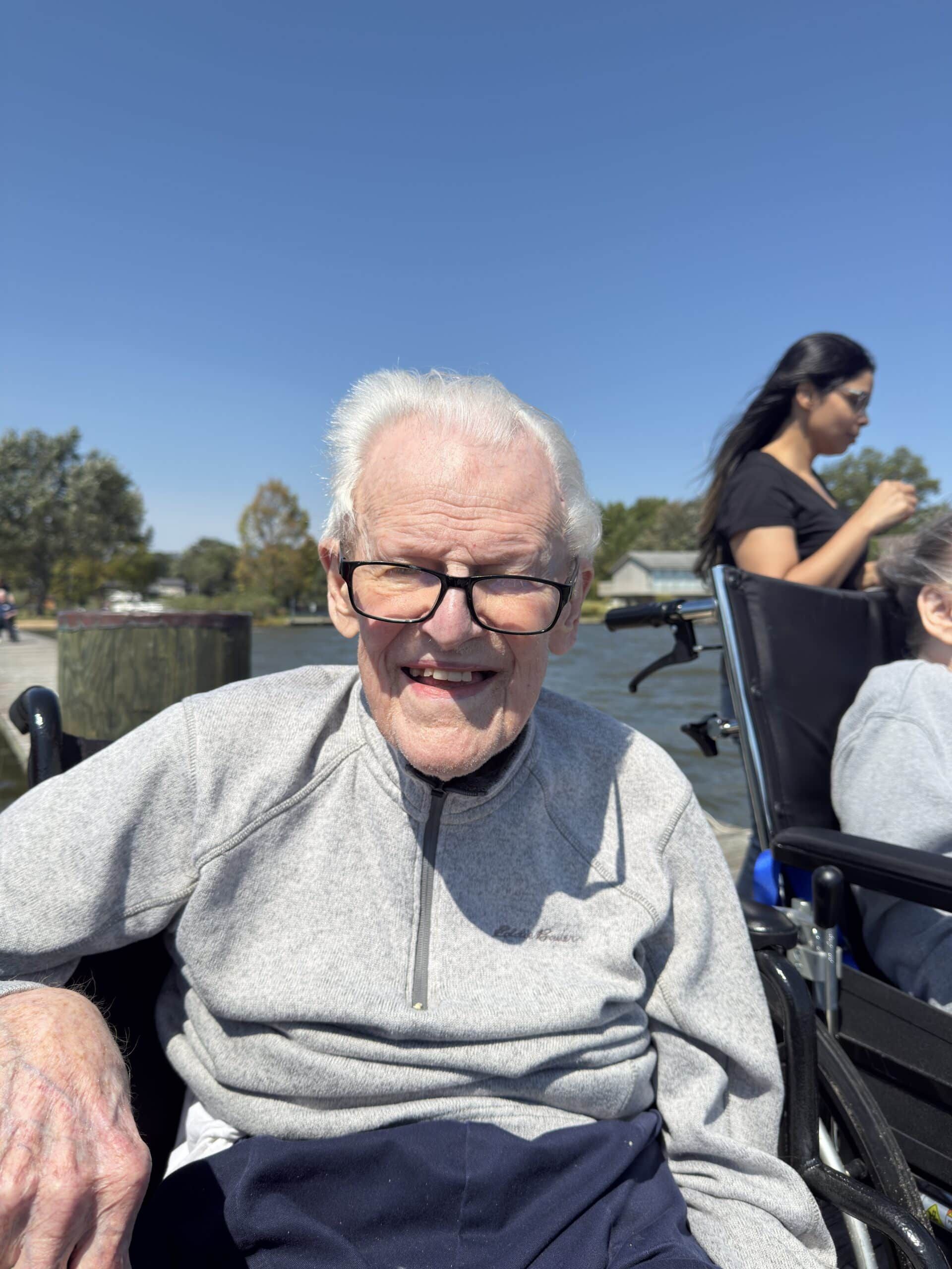A smiling person wearing glasses and a gray sweater in a wheelchair outdoors by a body of water.