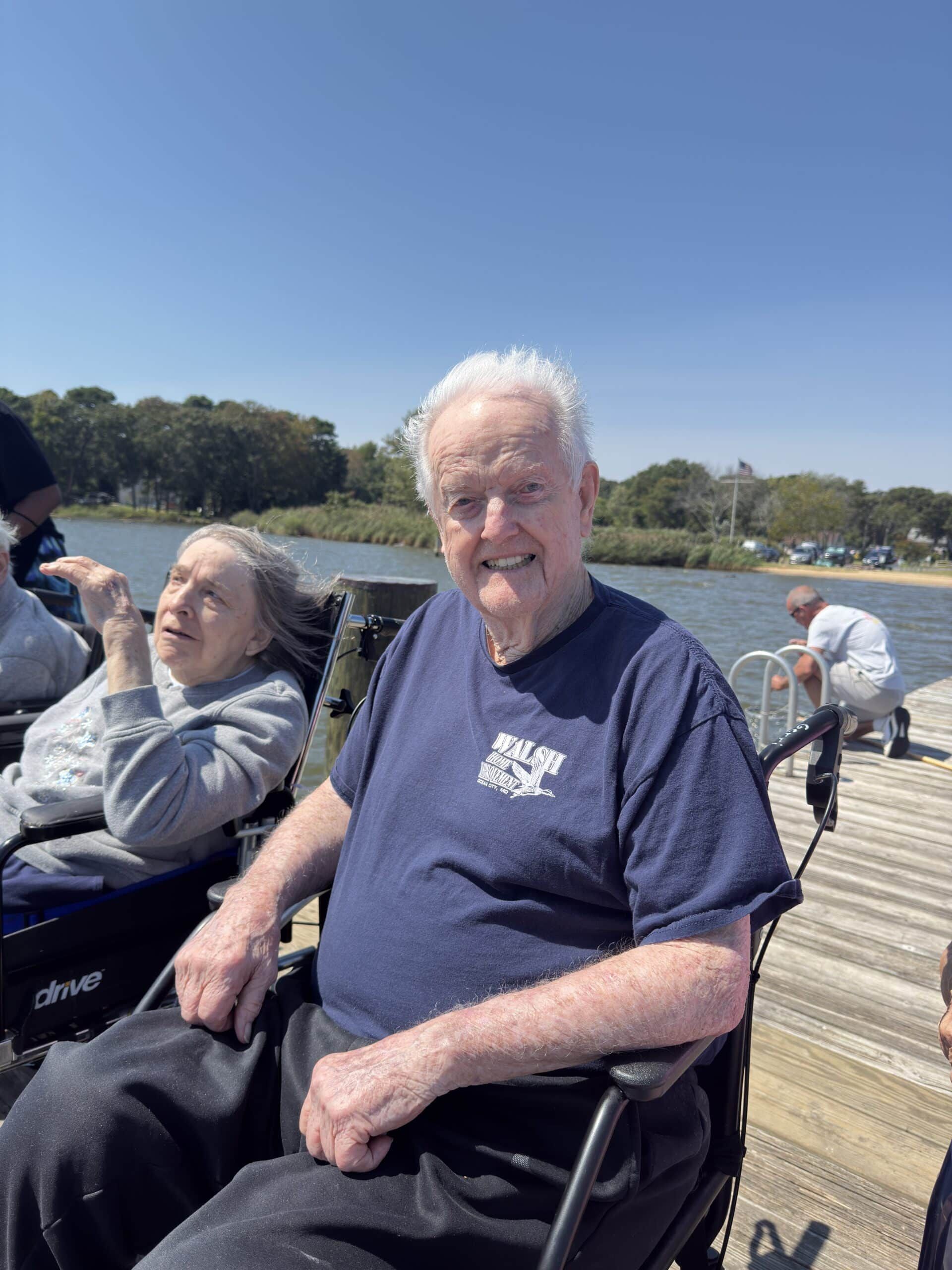 Two people in wheelchairs sit on a wooden pier near a lake on a sunny day, with a third person visible in the background.