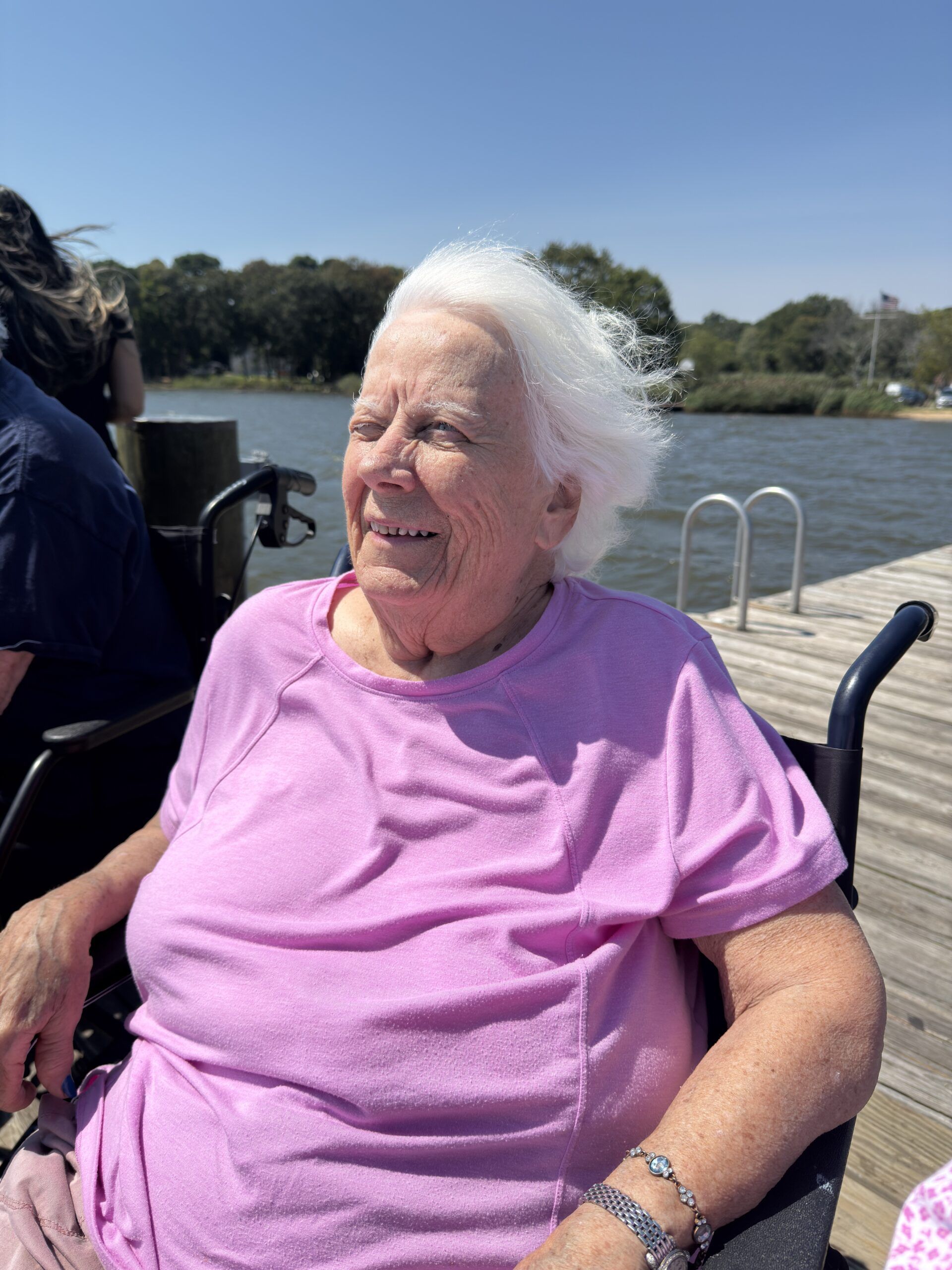A person in a pink shirt smiling while sitting in a wheelchair on a wooden dock overlooking a lake.