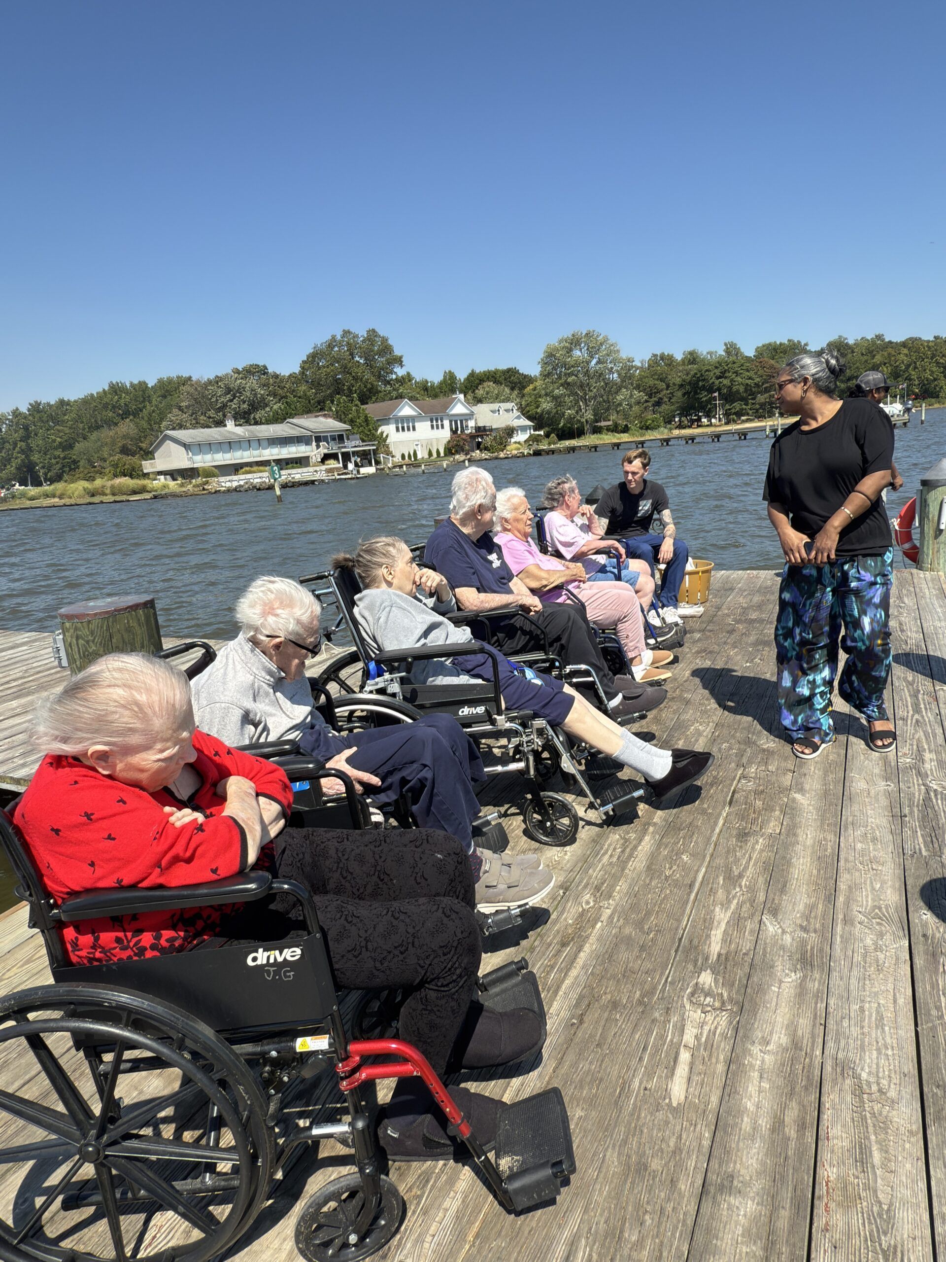 A group of people in wheelchairs sits on a wooden dock by a river, listening to a person standing nearby.