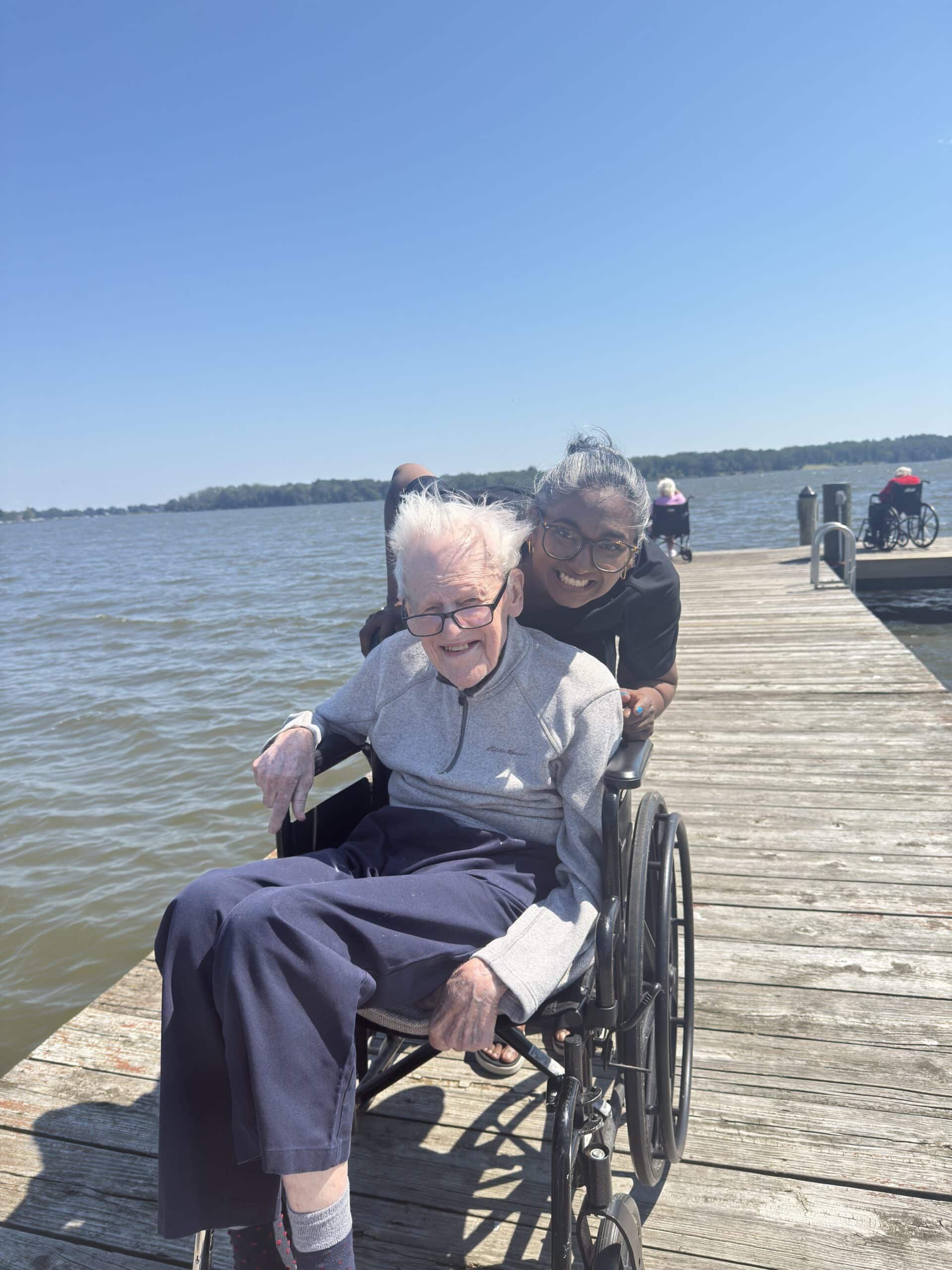 Two people, one in a wheelchair, smiling on a wooden pier overlooking a wide body of water under a clear blue sky.