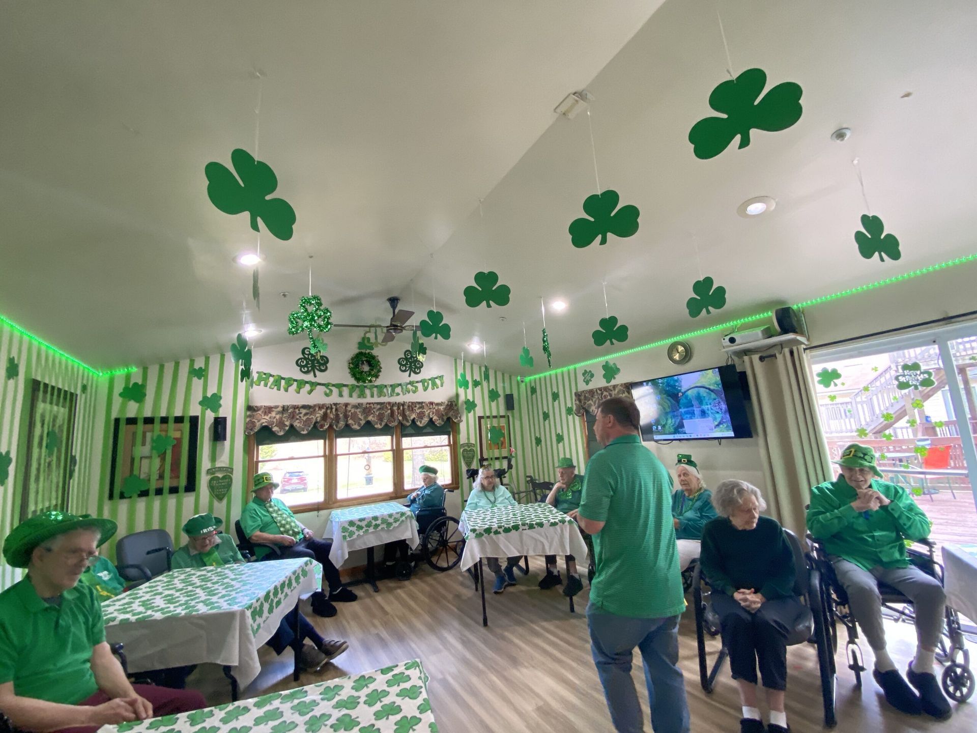 People in green clothing celebrate St. Patrick's Day in a room decorated with hanging shamrocks and festive banners.