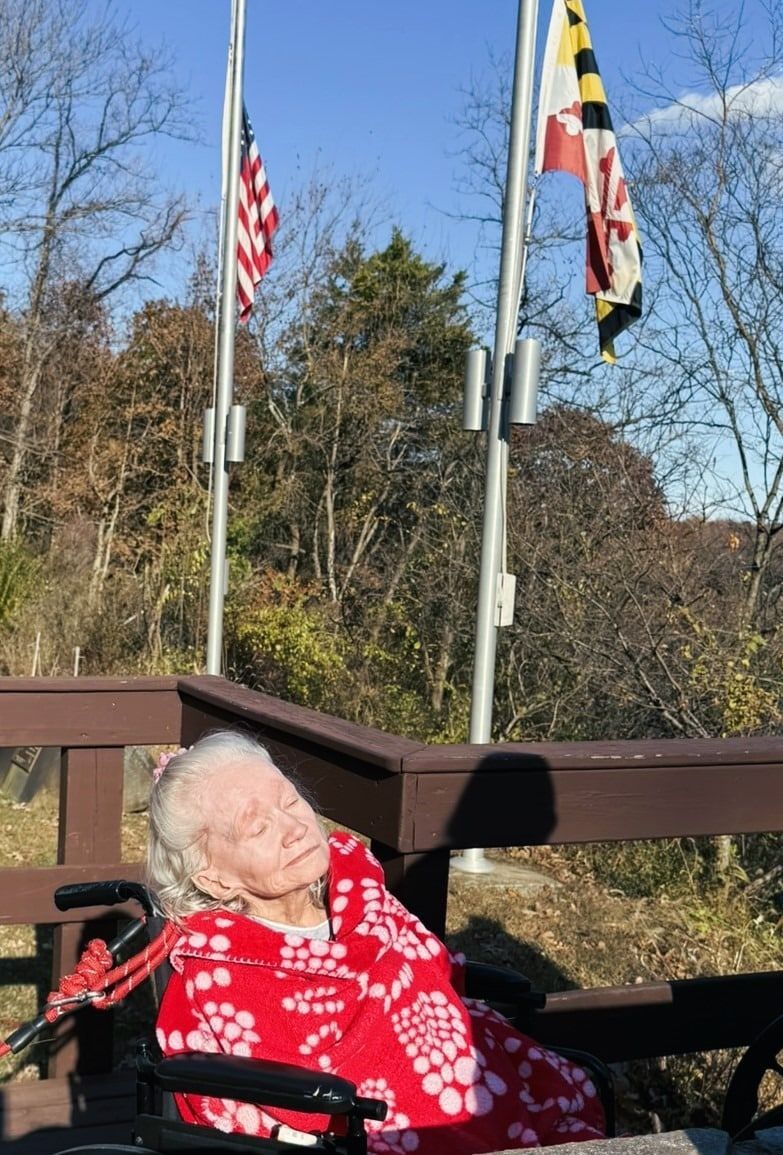 An elderly person with white hair, wearing a red patterned blanket, sits in a wheelchair outdoors beneath two flagpoles.