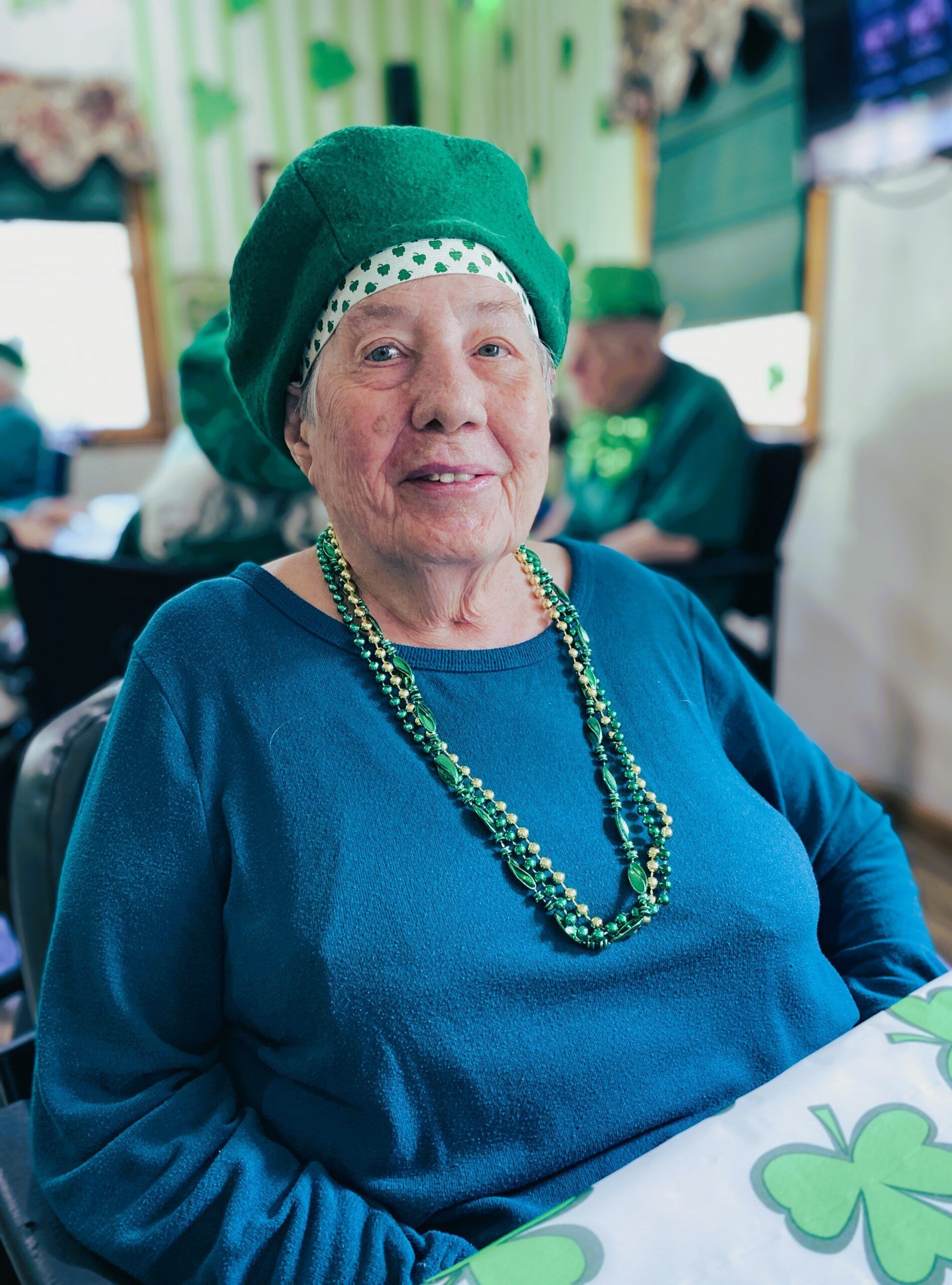 An older person wearing a green beret and green beaded necklace, smiling at a St. Patrick’s Day themed table.