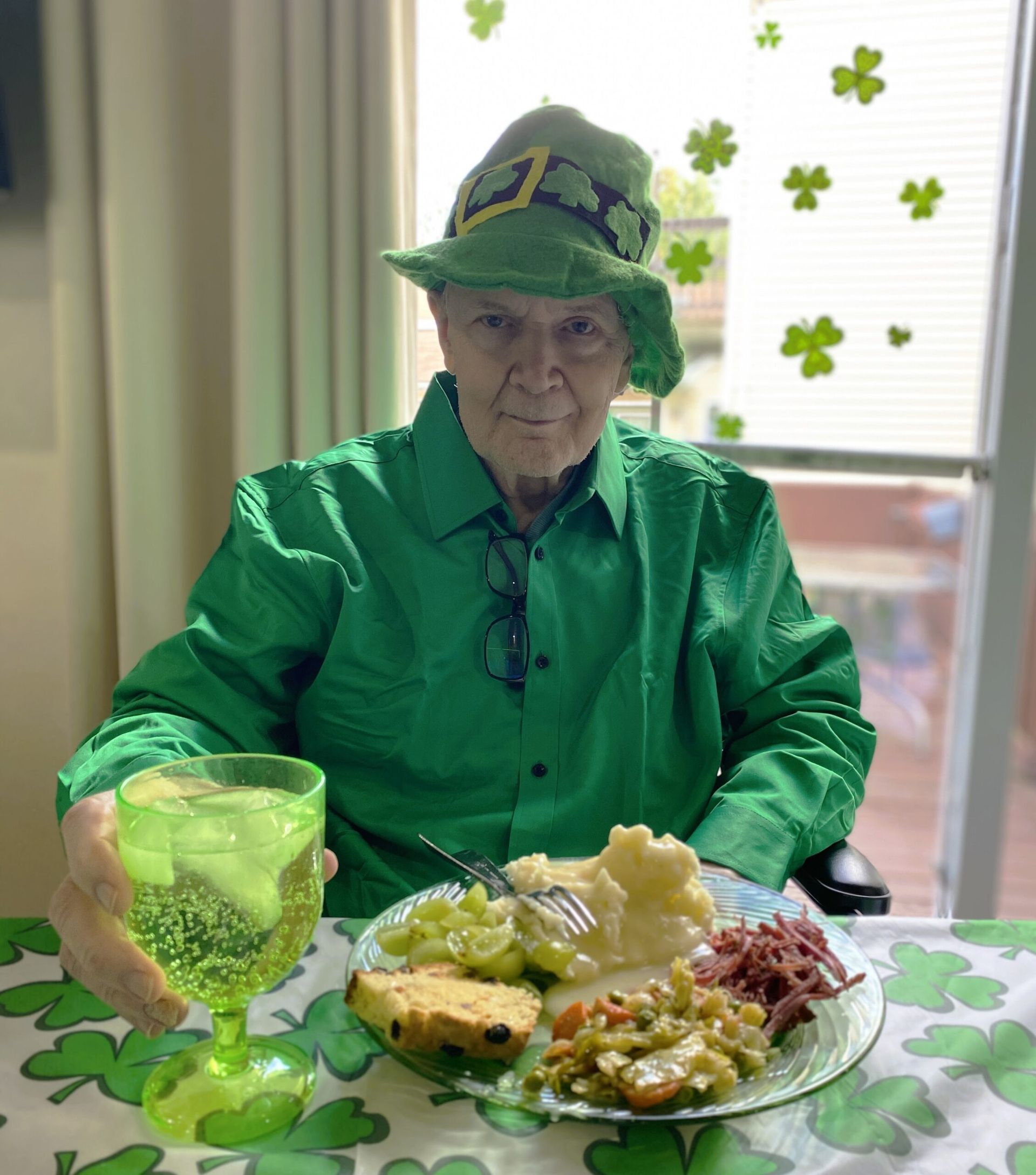 A person wearing a green leprechaun hat and shirt sits at a table with a meal and a green glass, celebrating St. Patrick's.
