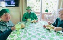 Three people wearing green celebrate St. Patrick's Day at a table with a shamrock tablecloth and themed food.