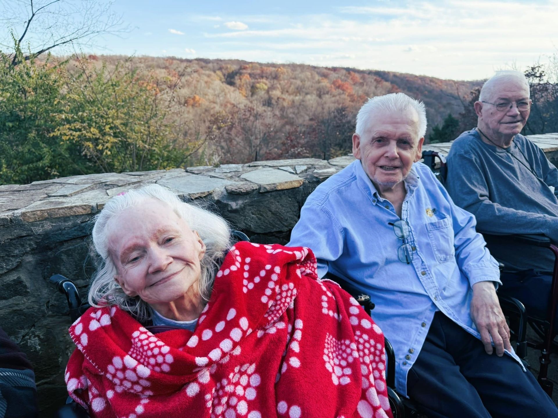 Three people sit outdoors on a stone wall overlooking a wooded landscape, with one wrapped in a red patterned blanket.