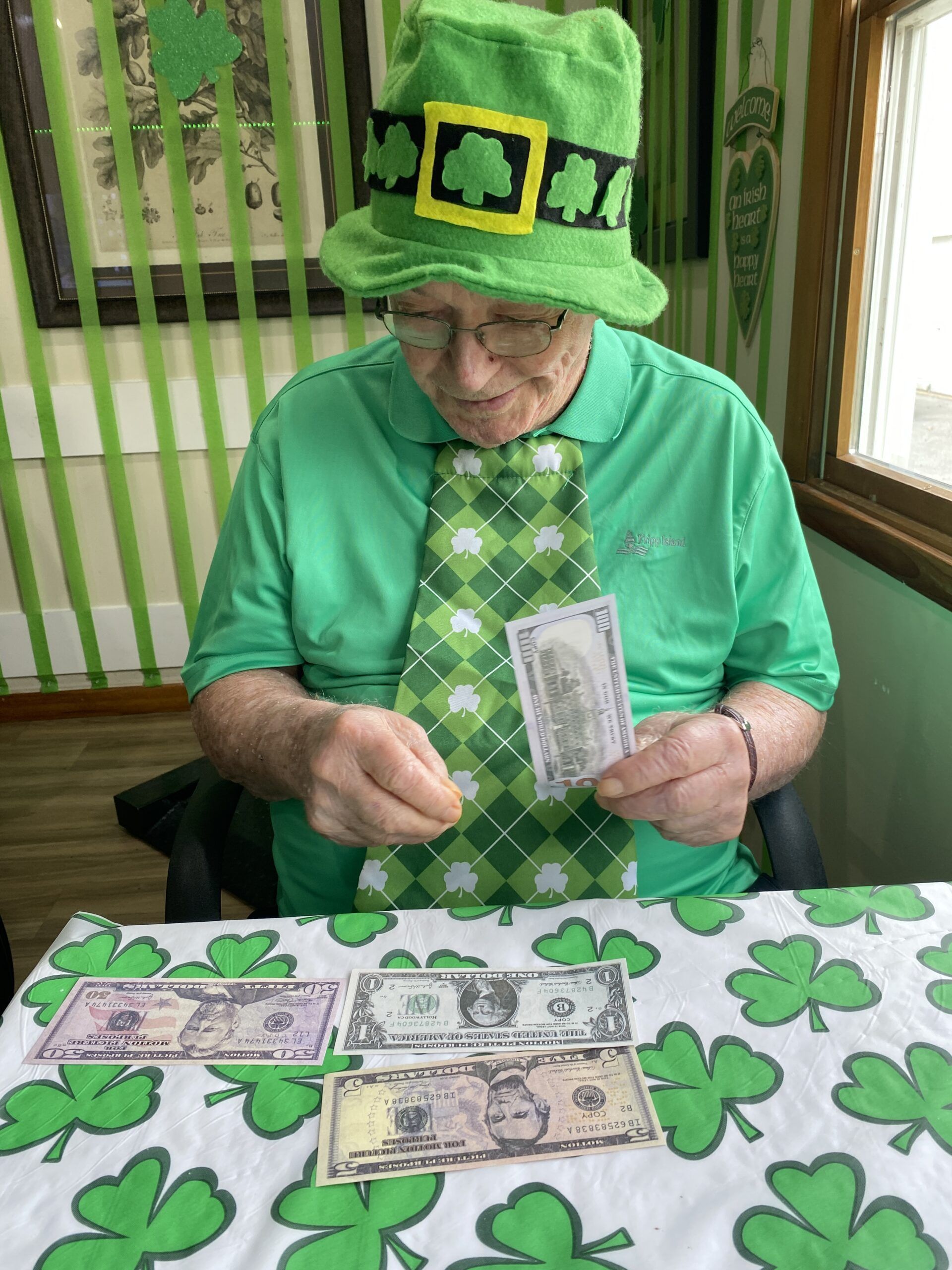 A person wearing a green shamrock hat and tie holds a bill while sitting at a table decorated with shamrocks and money.