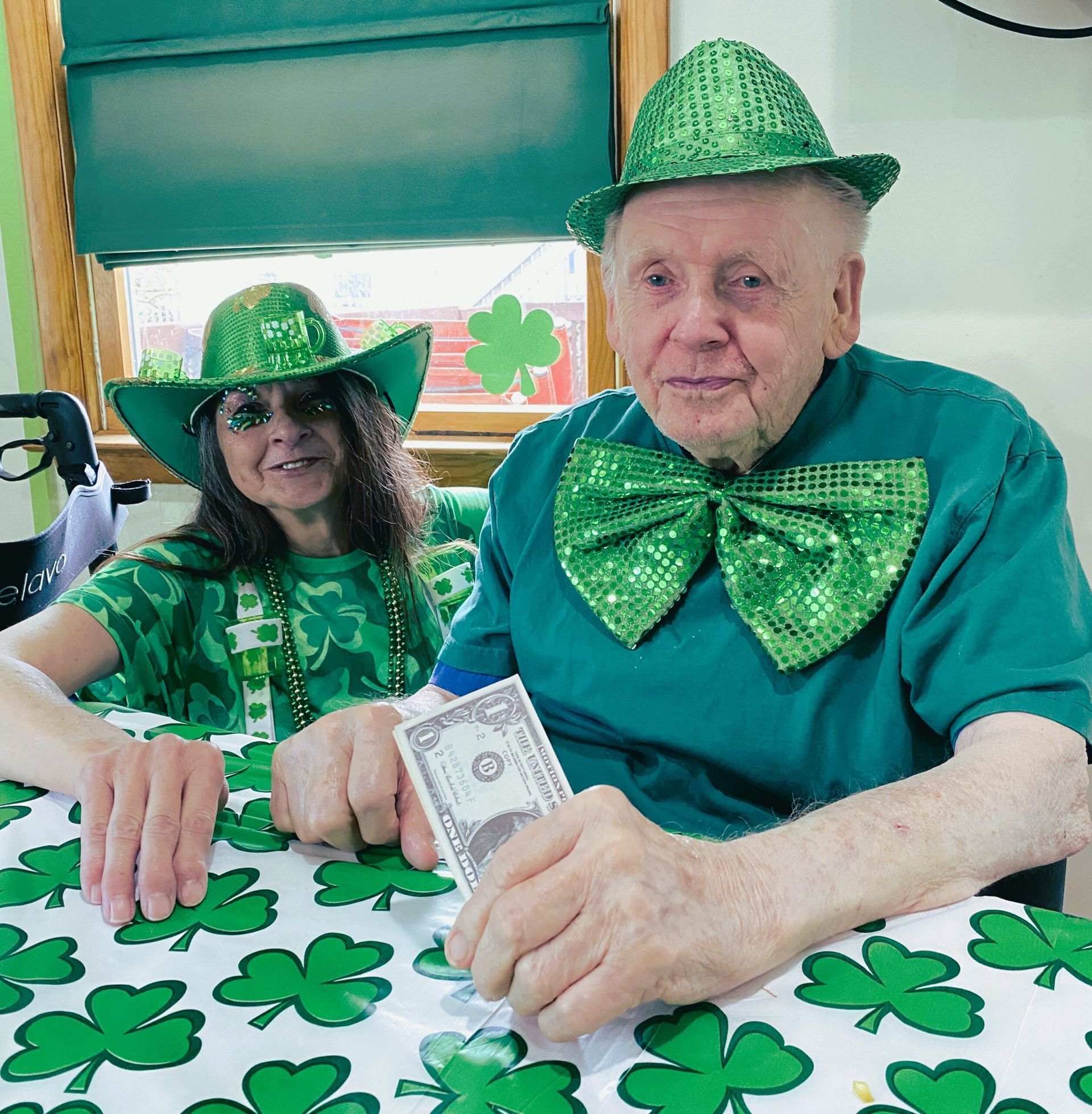 Two people in green hats and festive attire sitting at a table covered in a shamrock-patterned cloth holding a dollar bill.