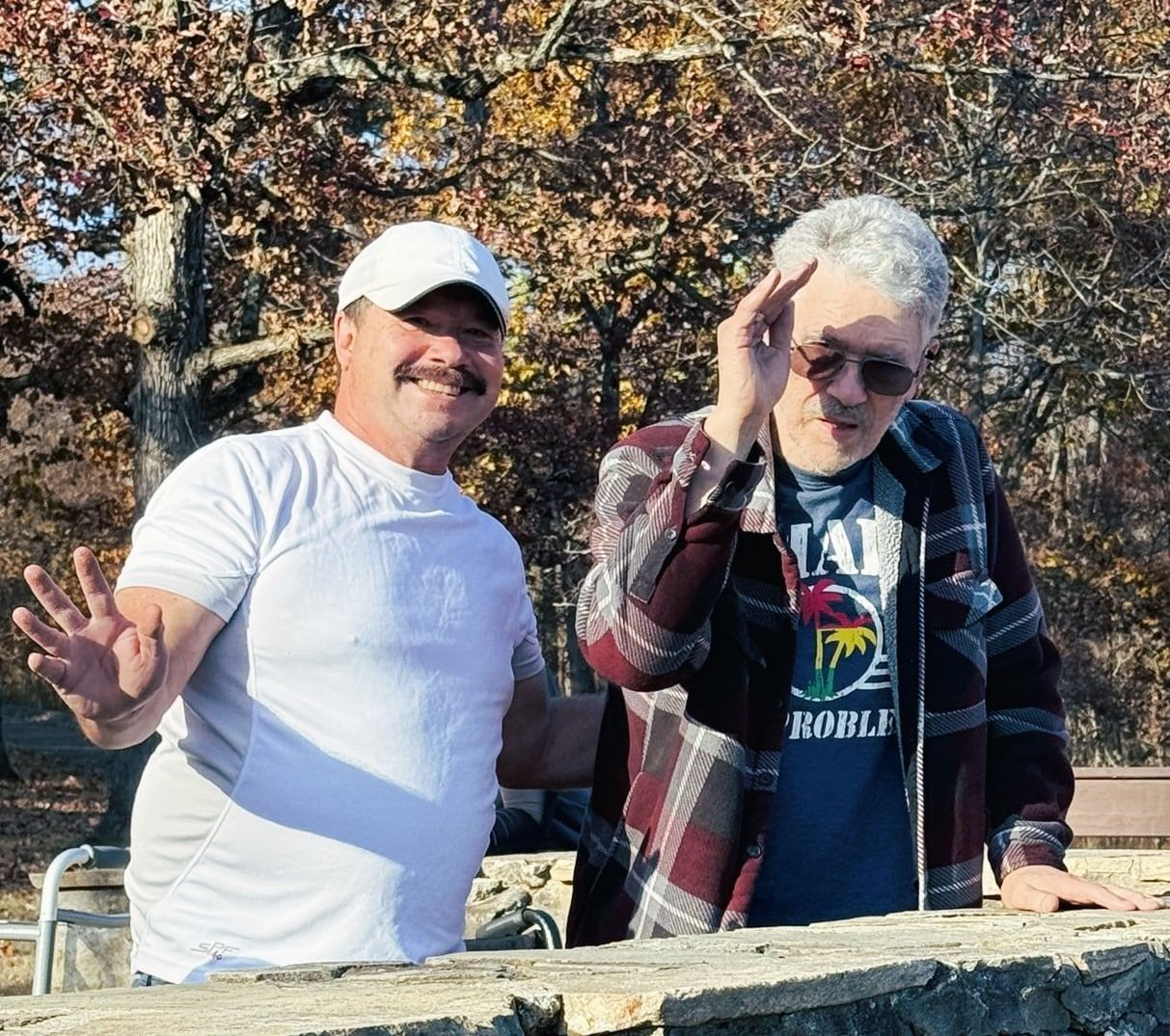 Two men standing outdoors in a wooded area, both waving at the camera. The person on the right wears sunglasses.