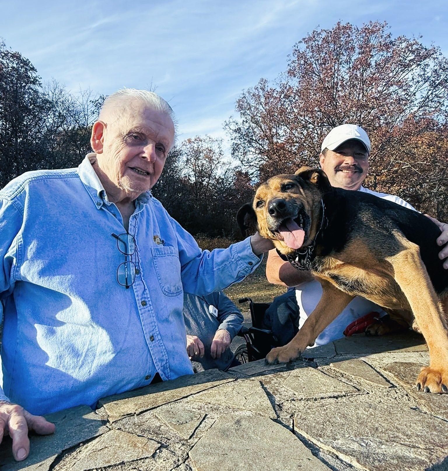 Two people stand outdoors with a smiling dog, positioned behind a stone wall with trees in the background.