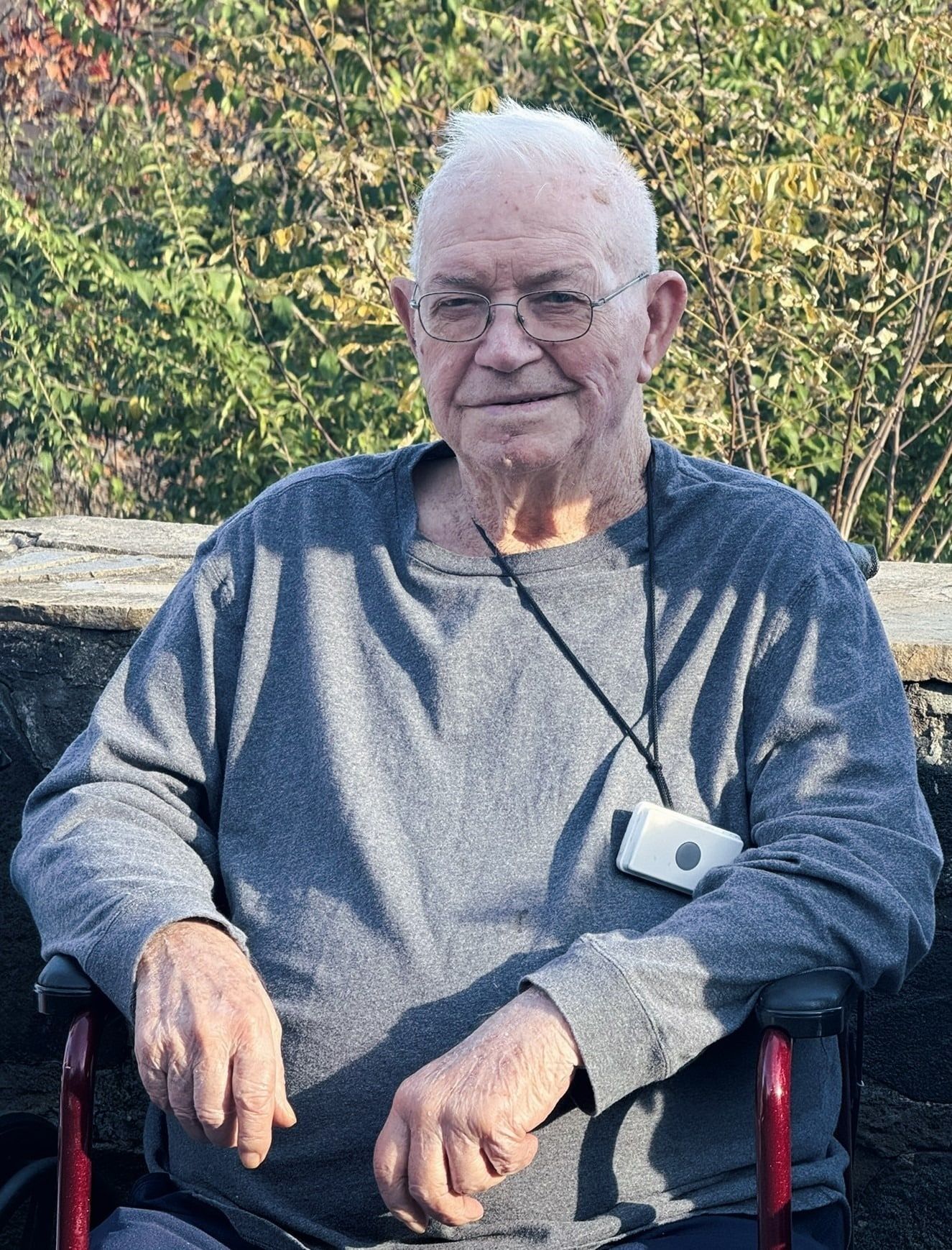An older person wearing glasses and a gray sweatshirt sits in a red wheelchair, smiling in front of a leafy background.