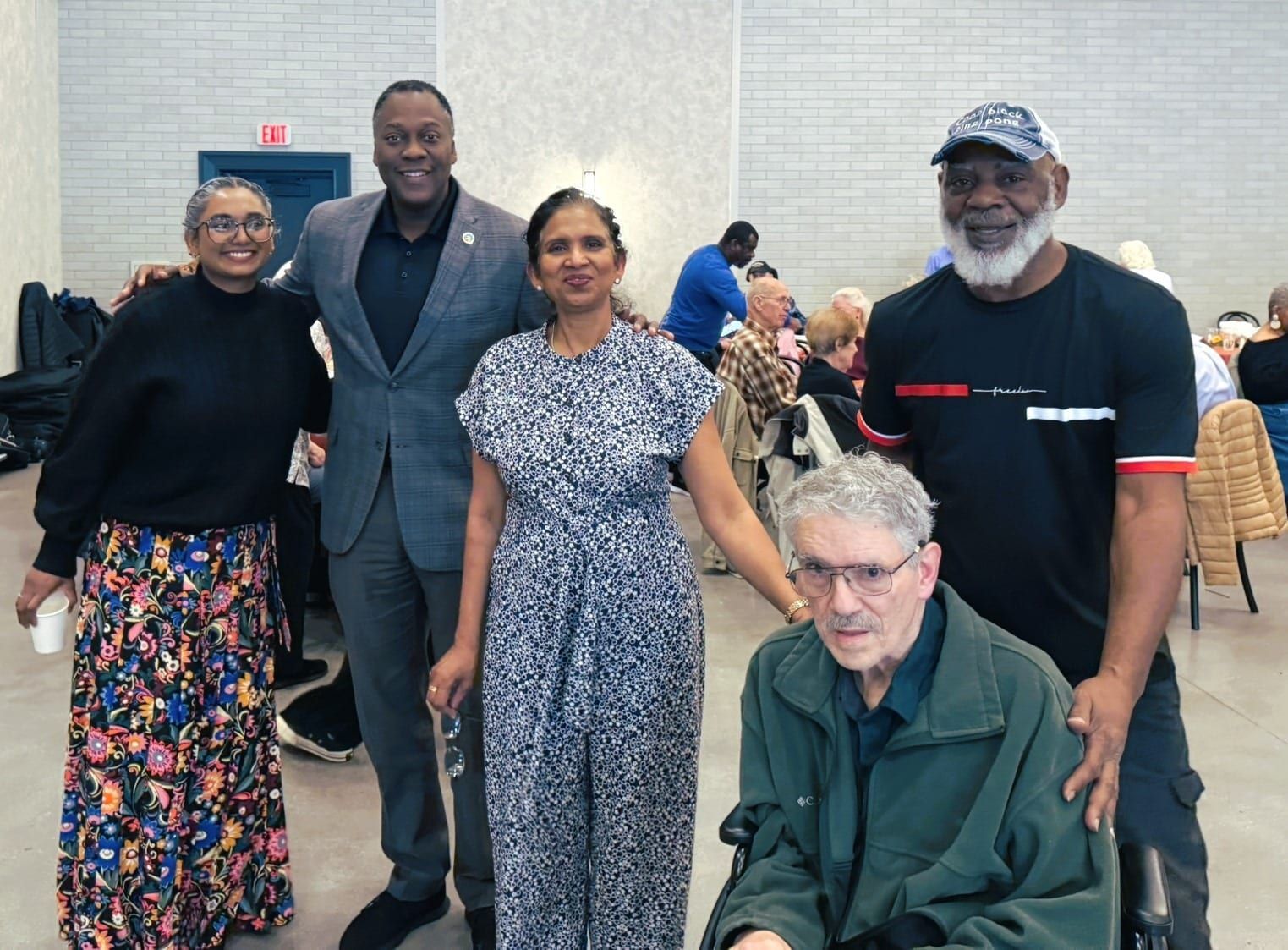 A group of five people stand and sit together in a large, neutrally toned indoor room, smiling for a group portrait.