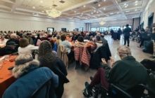 A crowd sits at tables in a large, bright banquet hall, facing an open area where people stand near the back.