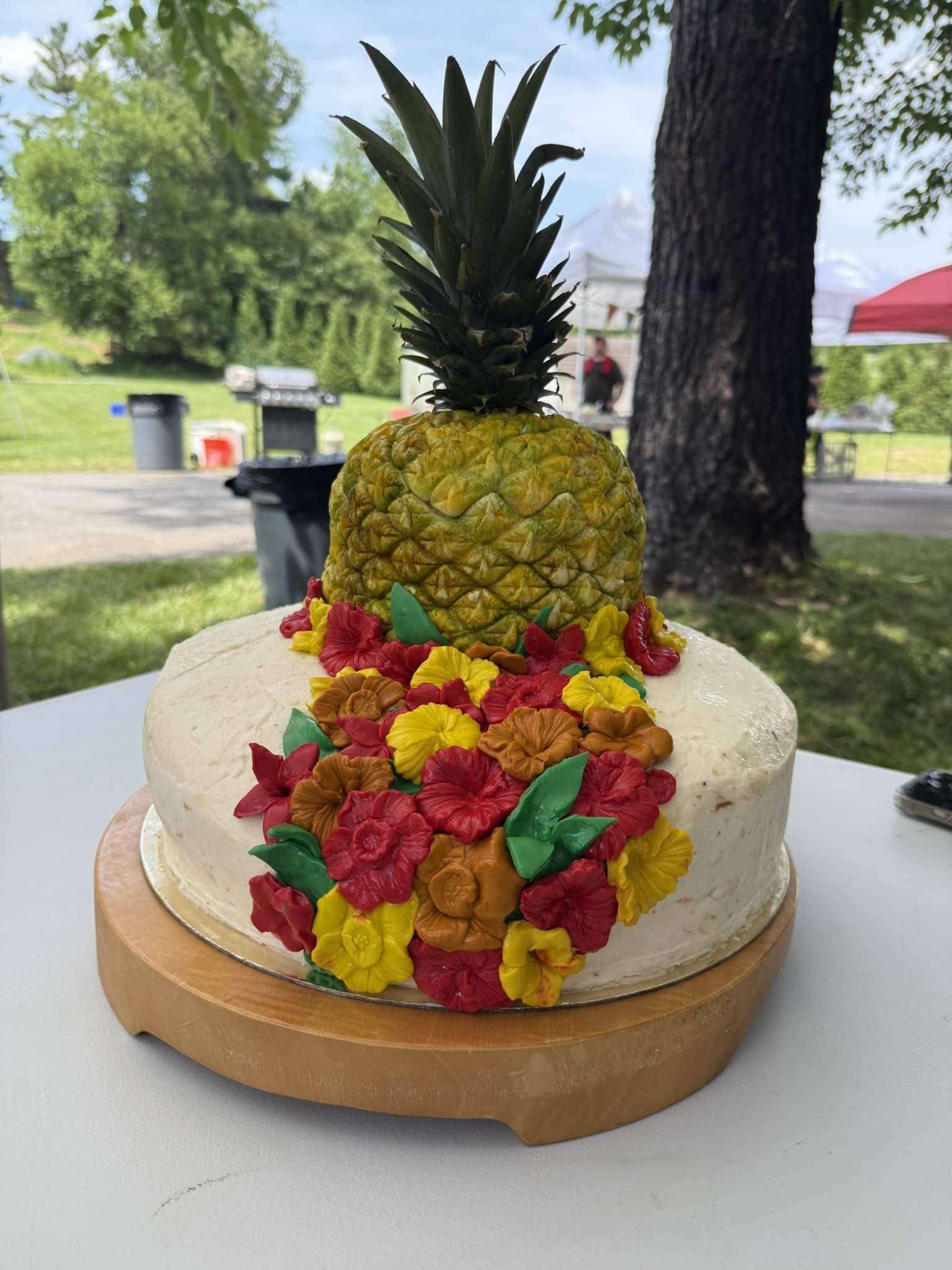 A pineapple-shaped cake topped with green fronds, resting on a round wooden board and decorated with colorful flowers.