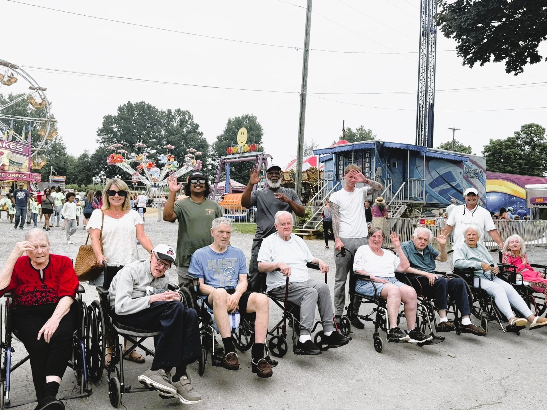 A group of people in wheelchairs and standing companions smile and wave at a fairground with rides in the background.