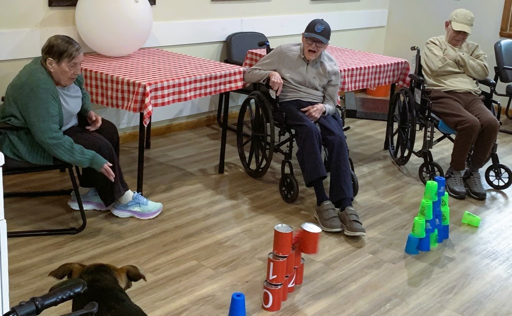 Three people in a room with red-checkered tables play a stacking game with red and blue cups, observed by a small dog.