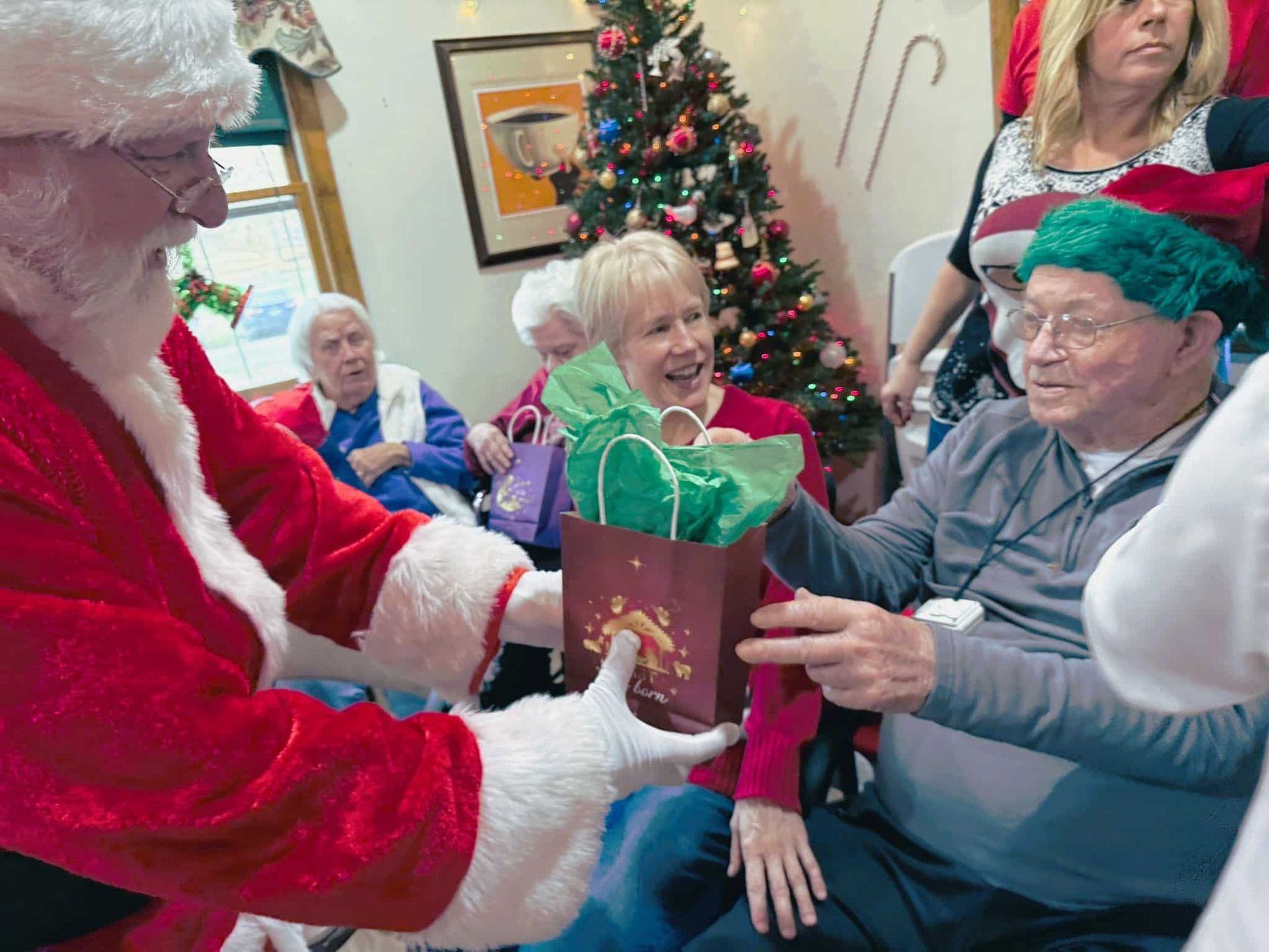 Santa Claus hands a gift in a green bag to an older person wearing a festive hat in a room decorated with a Christmas tree.
