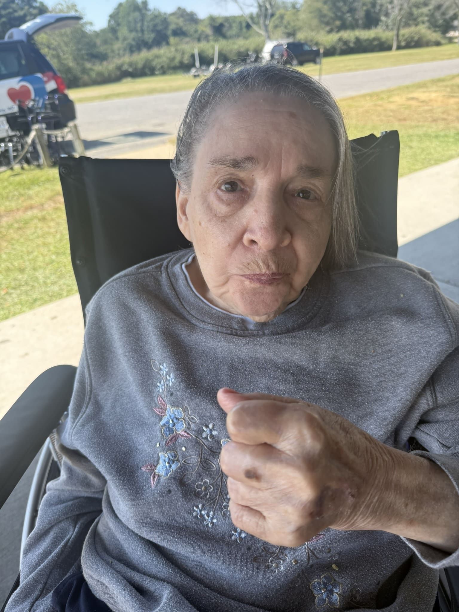 An elderly person in a gray sweatshirt sits in a wheelchair outdoors, holding their right hand in a closed fist.