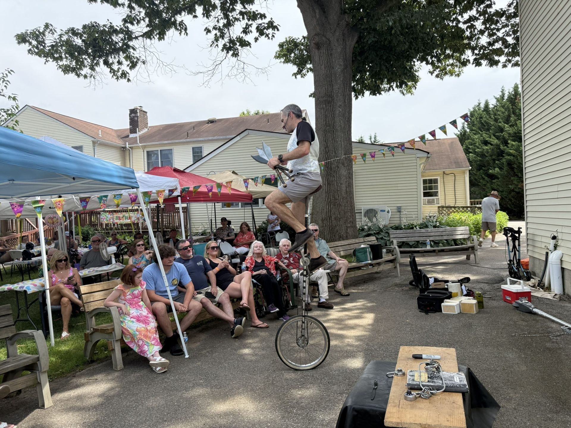 A person balances on a tall unicycle outdoors, entertaining a group of seated onlookers under a blue canopy and trees.