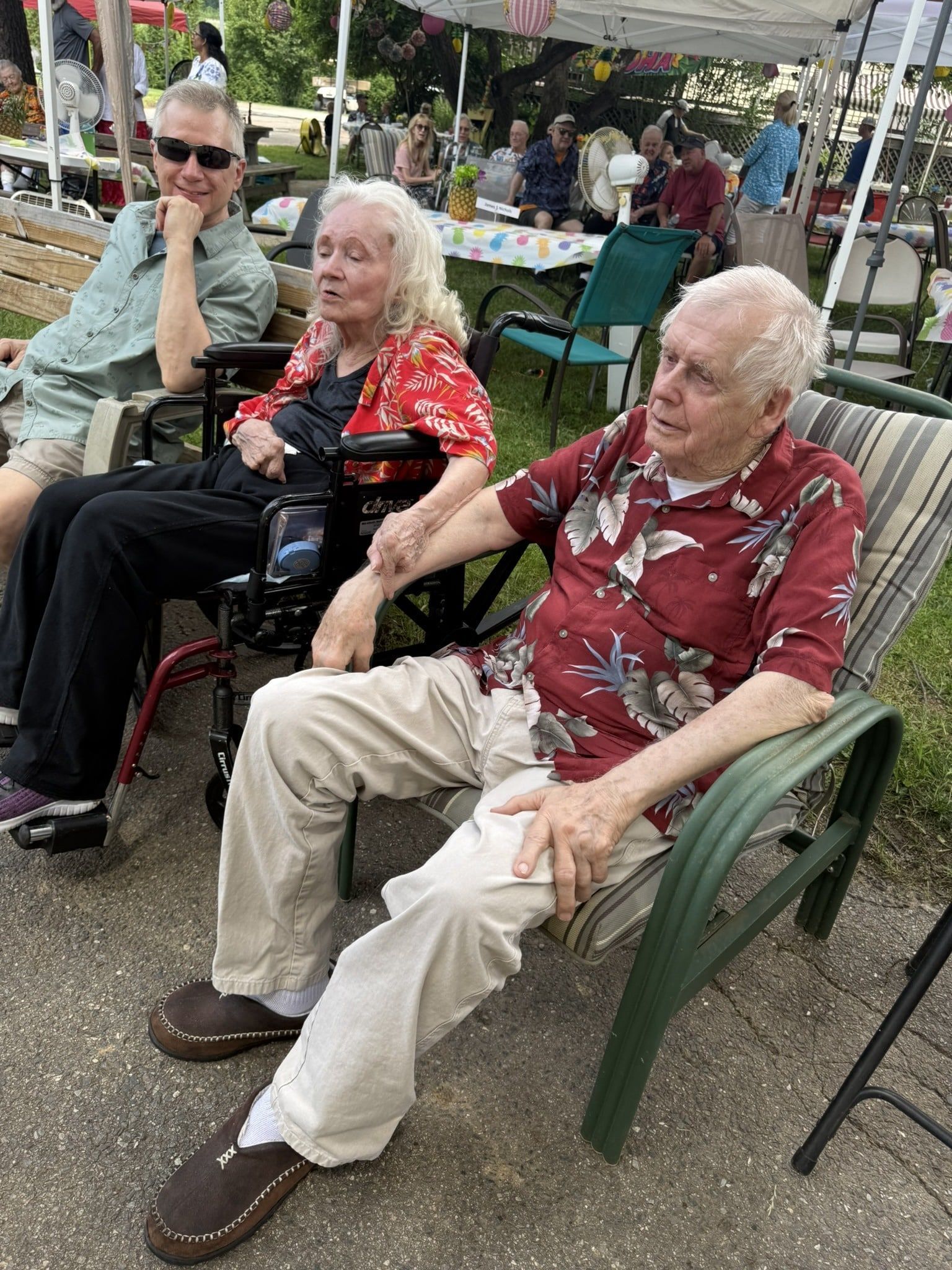Three people sit outdoors in lawn chairs, two wearing patterned shirts, at a sunny event with tables and shade canopies.