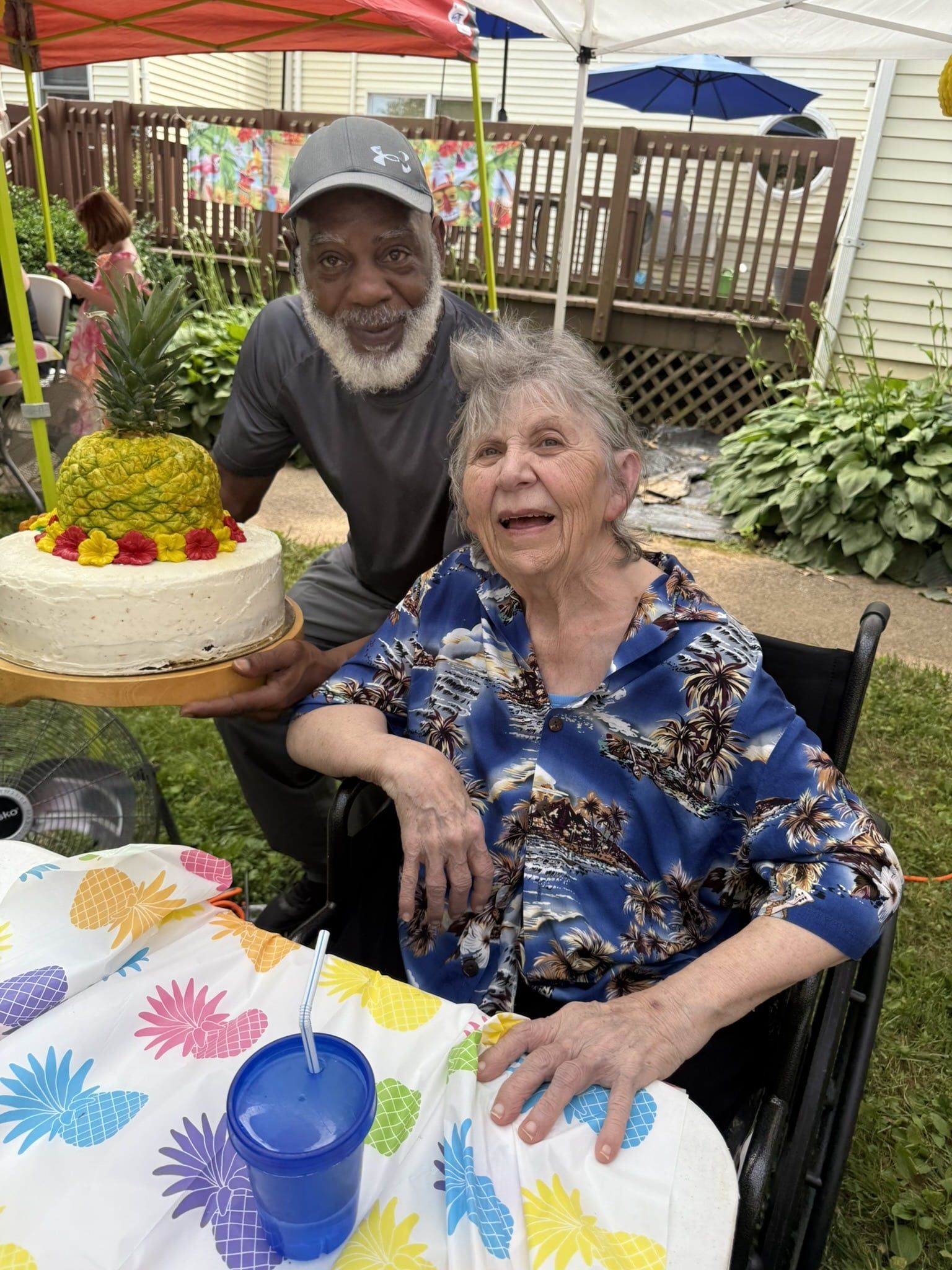An older person in a wheelchair smiles next to a person holding a cake topped with a pineapple at an outdoor celebration.