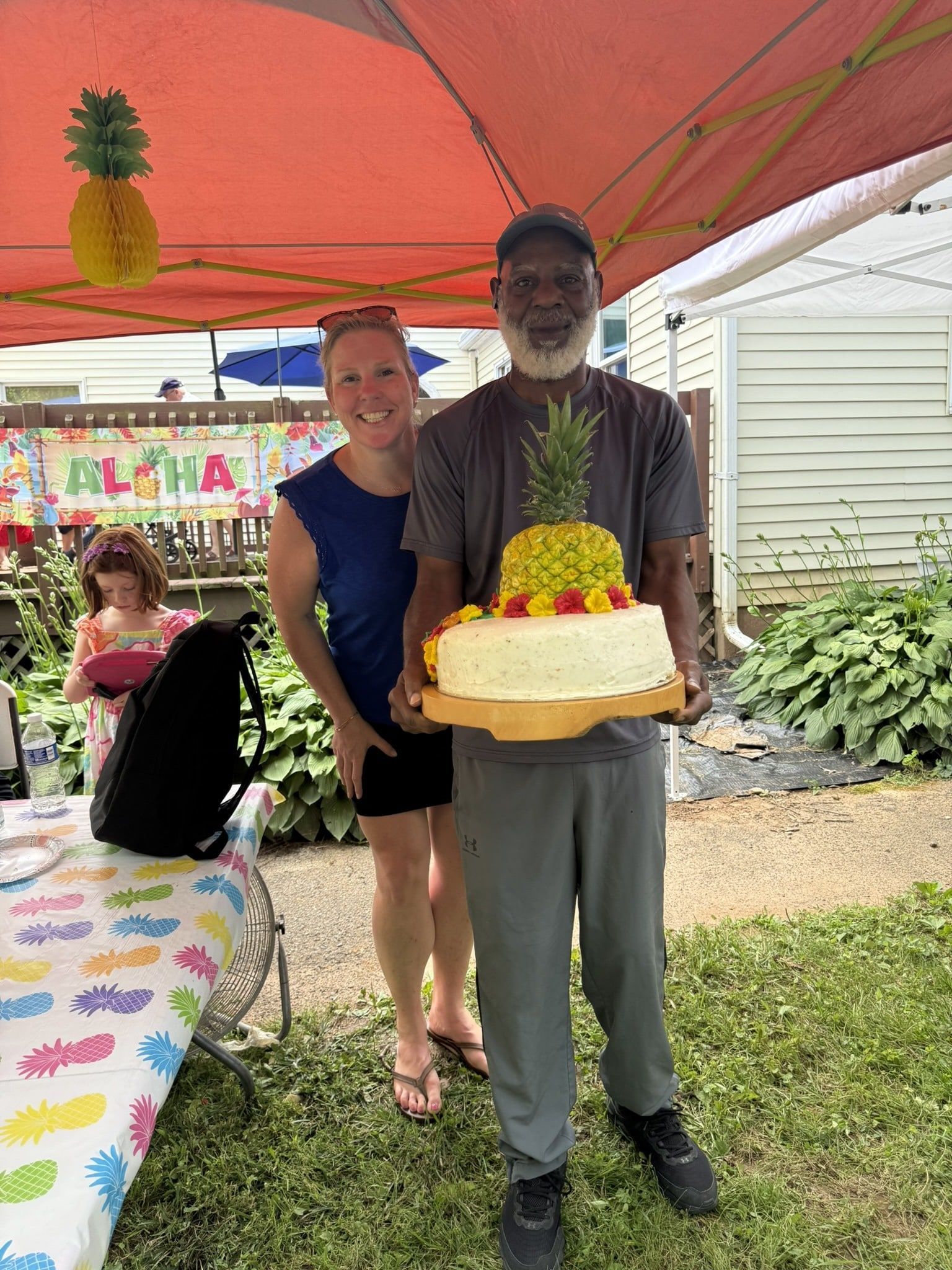 A person holds a pineapple-topped cake while standing next to another person outdoors under a tent at a party.