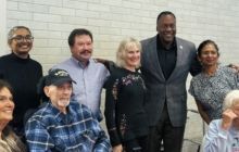 A group of eight people smiling for a photo indoors in front of a white brick wall.