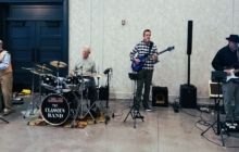 A four-piece band performs on a stage with a drum set, guitars, and audio equipment in an indoor hall.