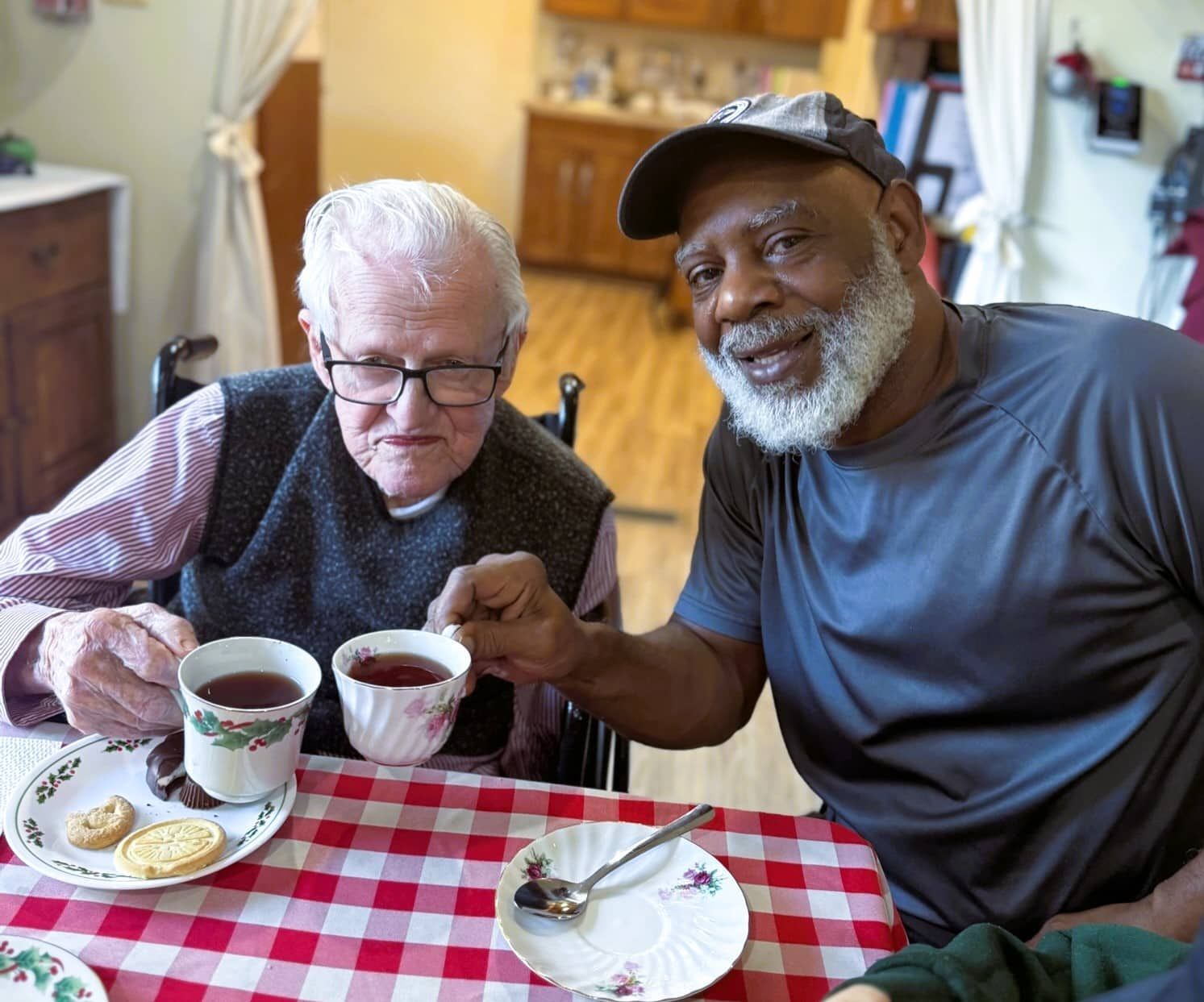 Two people sitting at a table with a red-and-white checkered cloth, clinking their tea cups together with smiles.