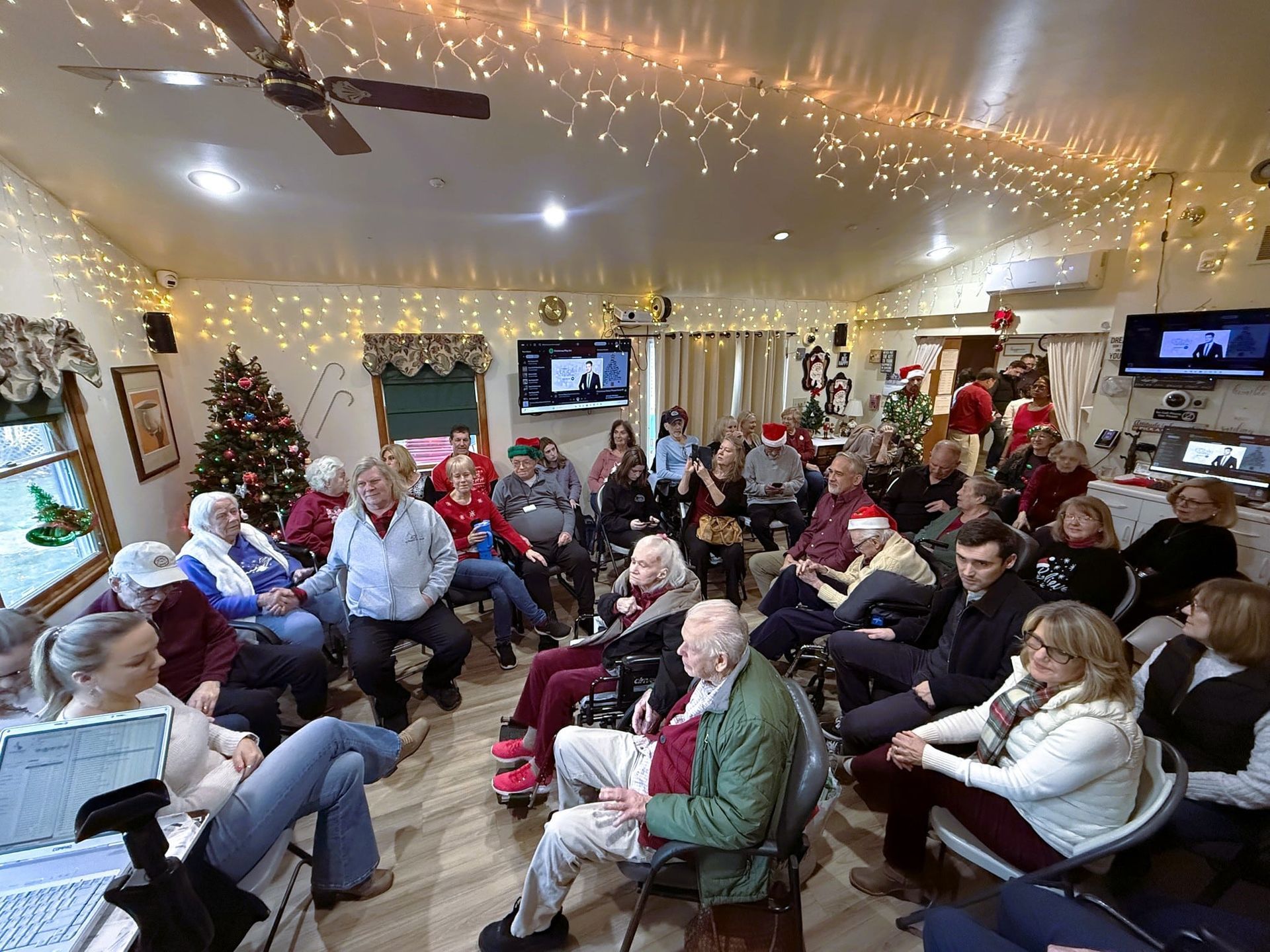 A group gathers in a decorated community room with string lights and a Christmas tree for a holiday activity or event.