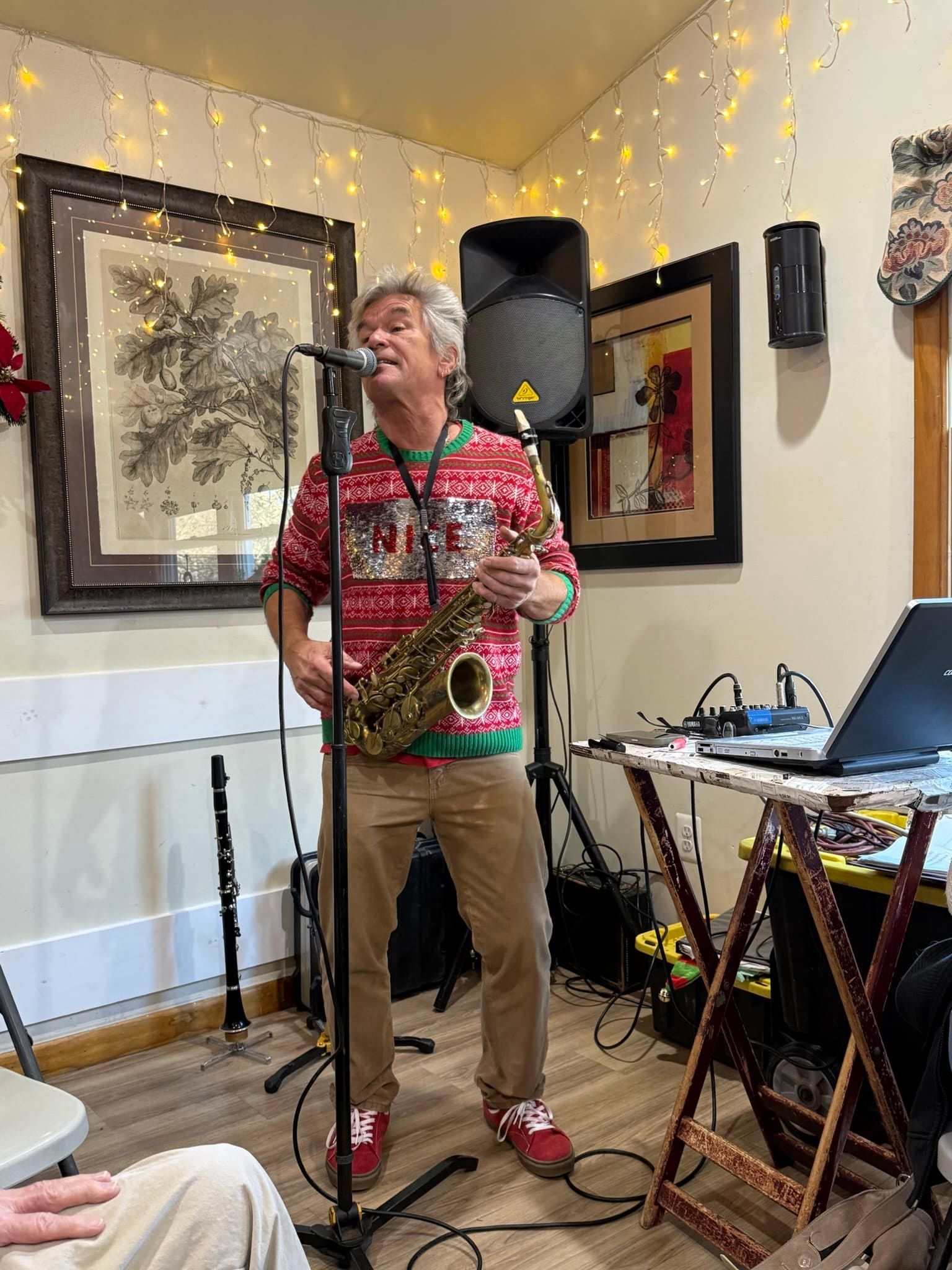 A musician in a festive sweater plays a saxophone while standing in front of a microphone and speakers.