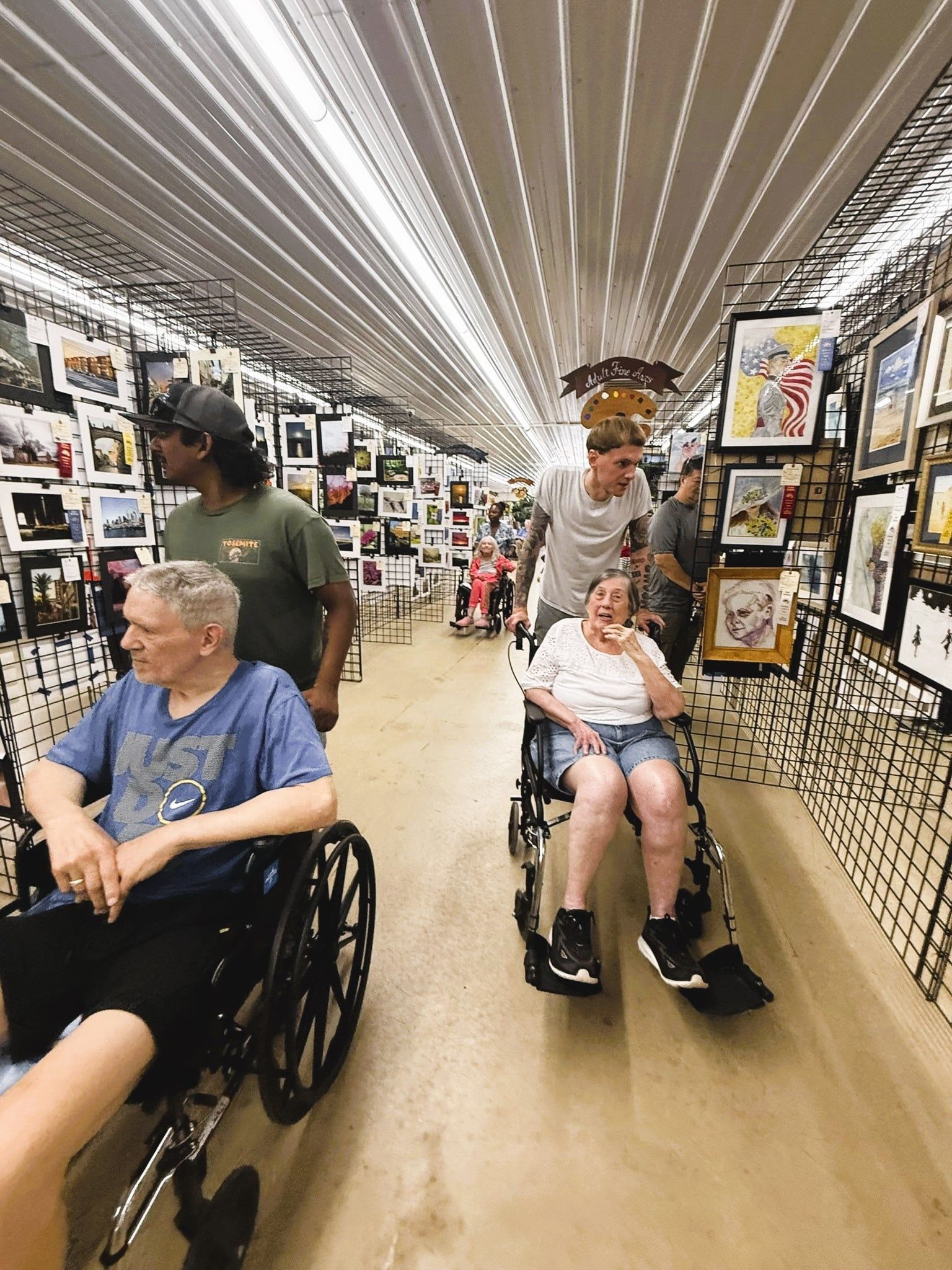 People in wheelchairs move through a hallway lined with framed artwork on both sides under bright fluorescent lighting.