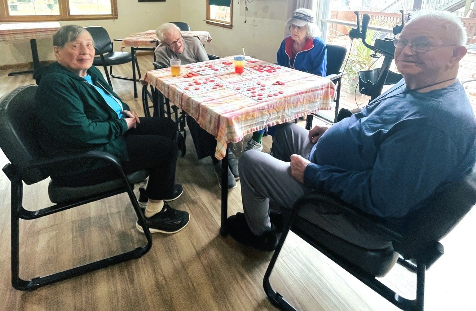 Four people sit around a table with a patterned cloth in a room with light wood floors, some looking toward the camera.