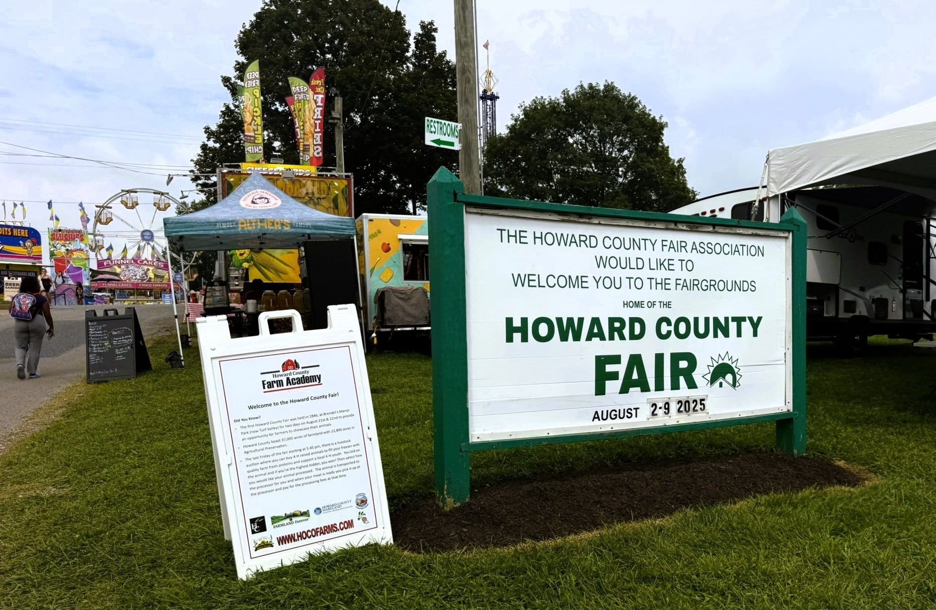 A sign for the Howard County Fair stands on grass in front of fairgrounds with food stands and a ferris wheel.