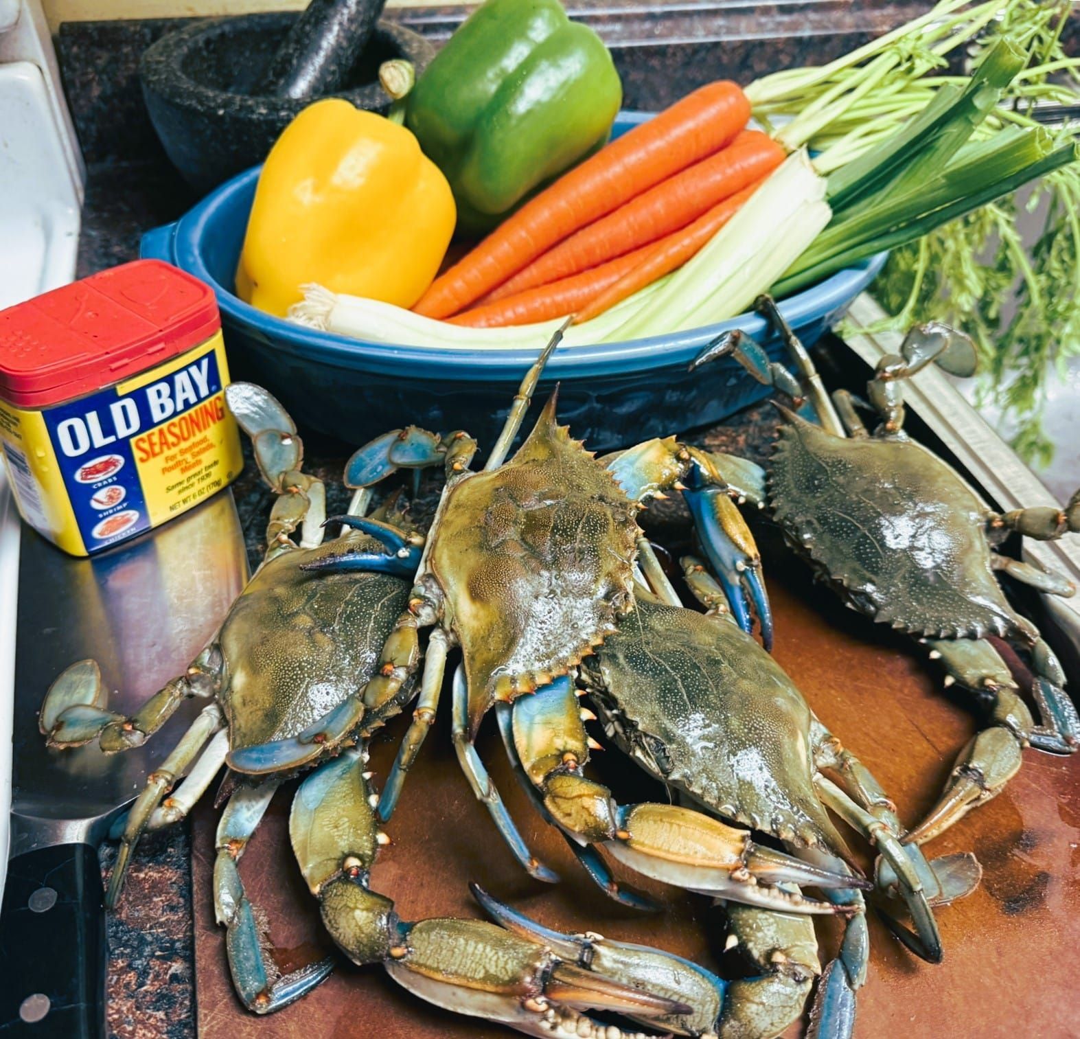 Blue crabs, a container of Old Bay seasoning, and a bowl of fresh vegetables on a kitchen counter.