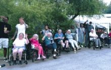 A group of people sitting and standing outdoors in front of trees and tents, many waving and smiling at the camera.